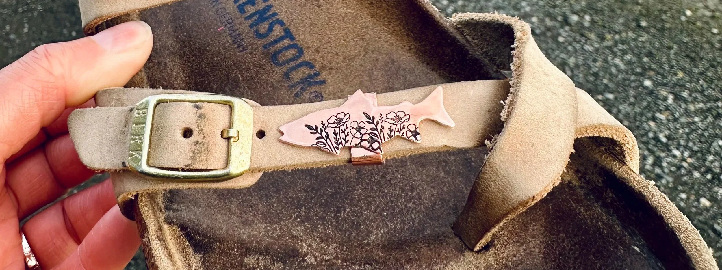Close-up of a beige suede Birkenstock sandal with a copper salmon-shaped charm decorated with black floral patterns. The background shows a beach setting.