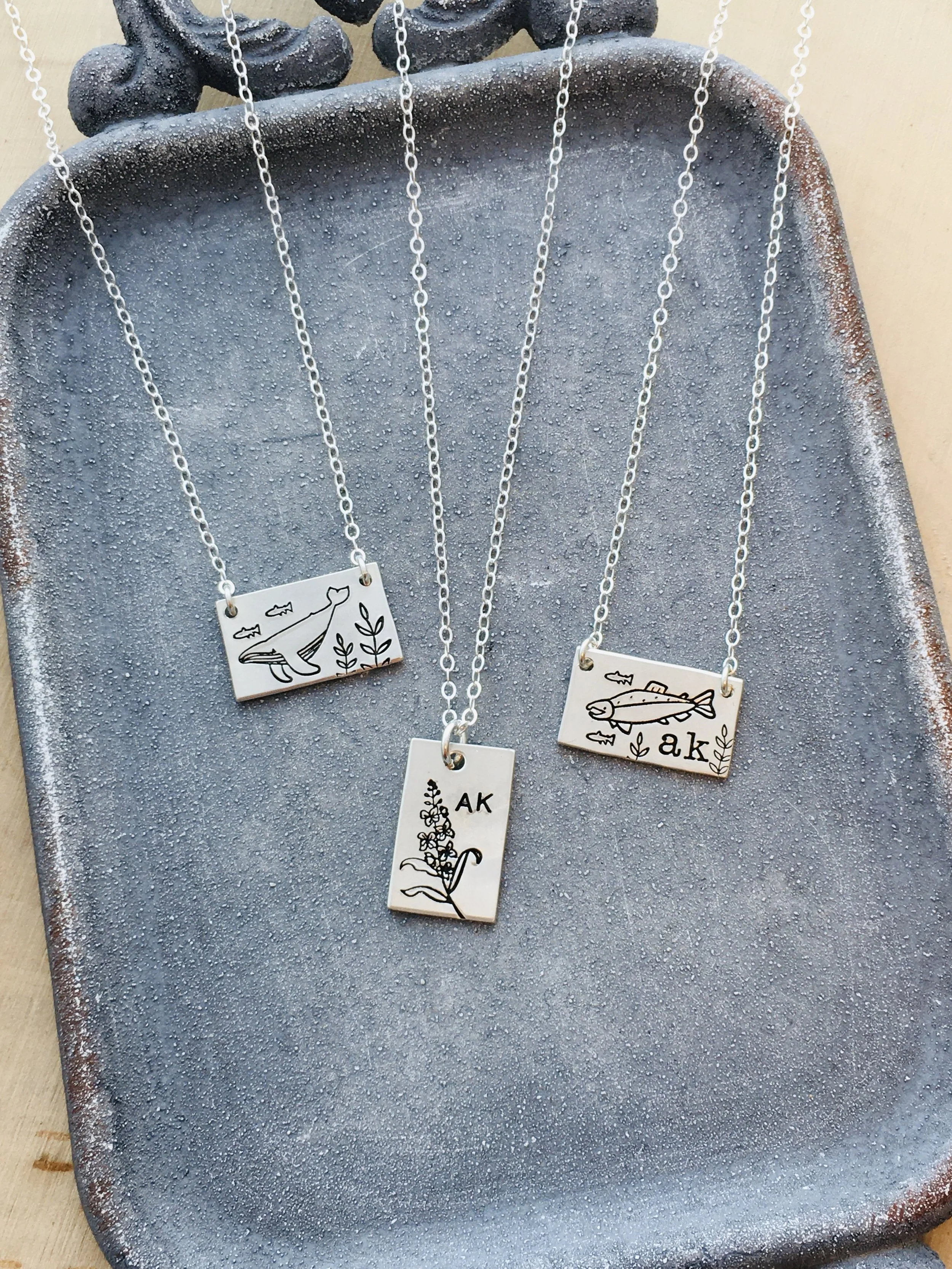 Sets of silver necklaces with rectangular pendants hanging on a textured, grayish-blue display. The pendants feature simple black line drawings of fish and plants, with initials 'a.k' on some.