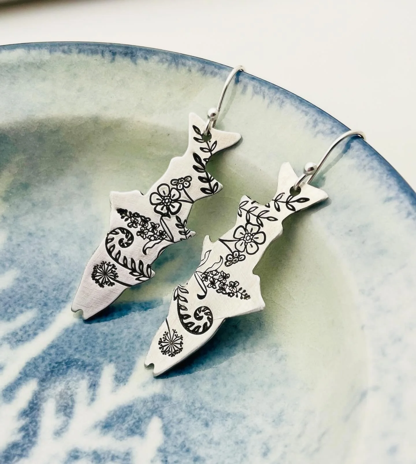 White earrings with black floral and vine designs hanging on a blue and green ceramic dish.