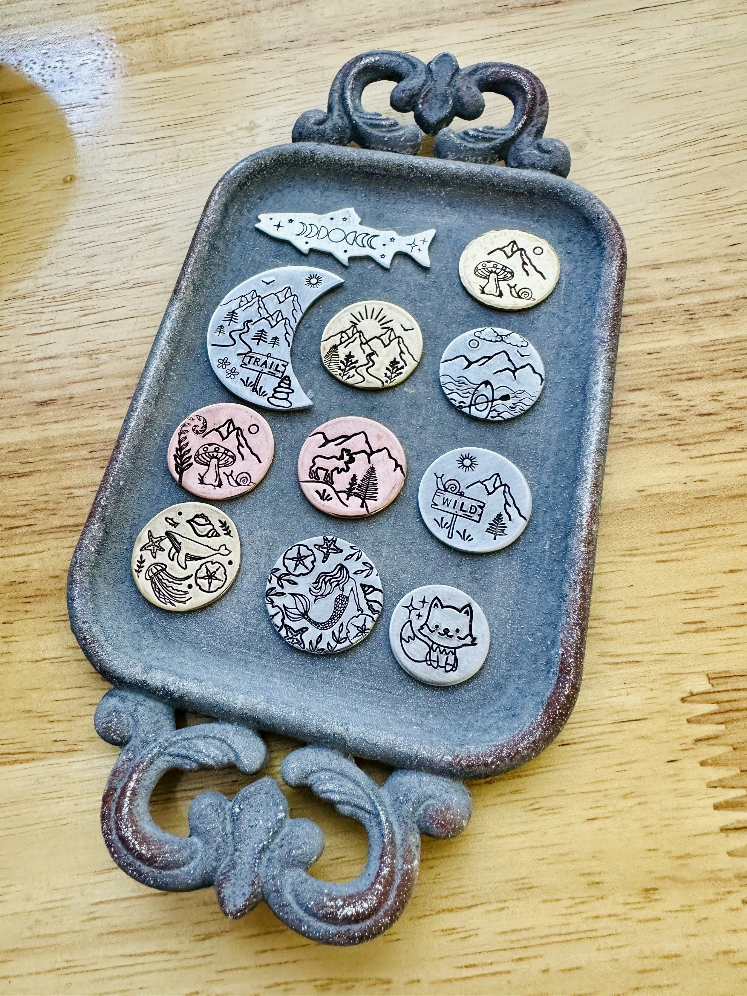 A vintage metal tray with decorative handles contains various pins with nature and mountain motifs, some featuring moon and sun designs, a fox, a whale, a fox, and a wild sign.