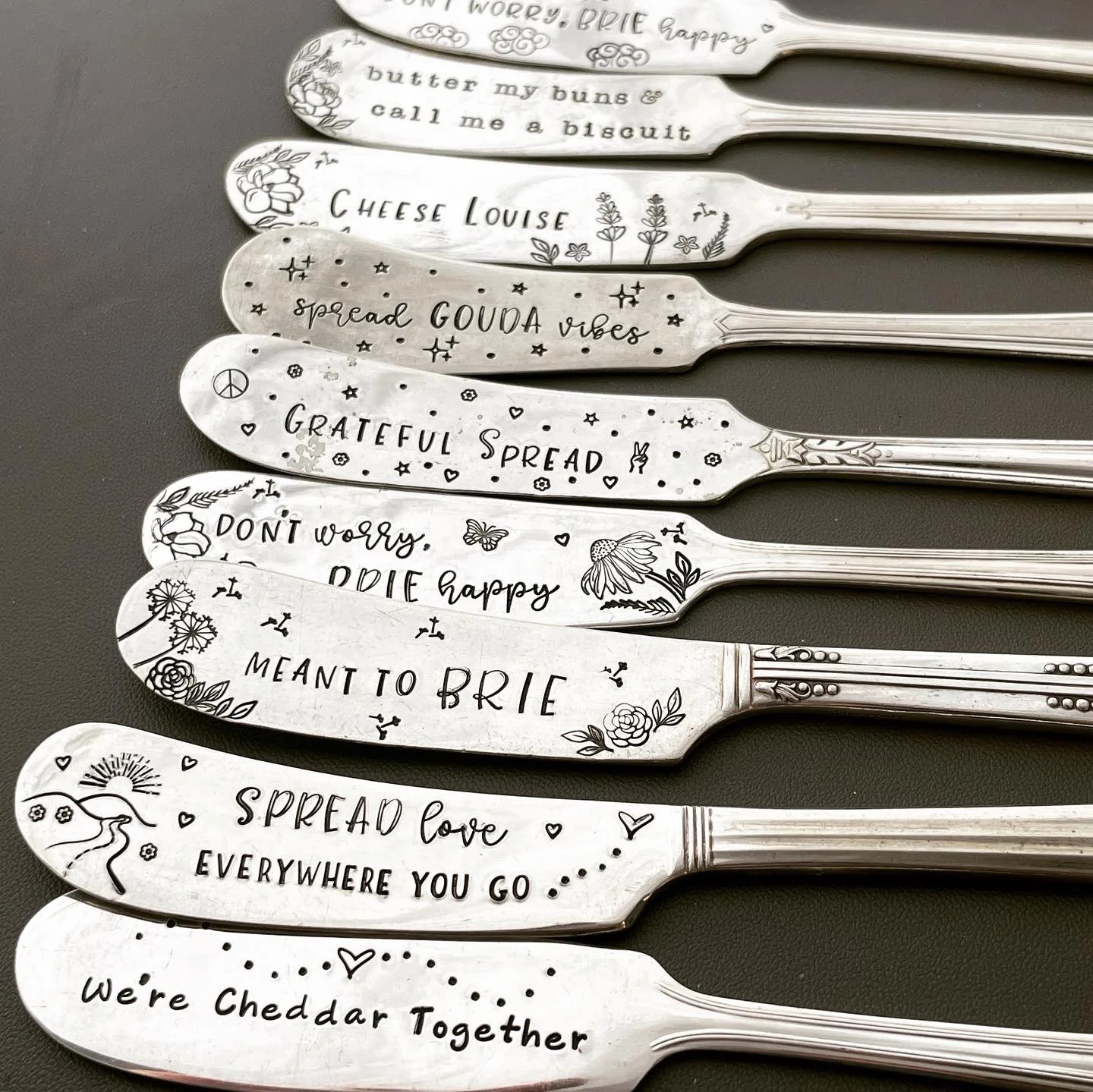 Collection of decorative silver butter knives with engraved phrases and drawings, each featuring different inspirational or humorous messages, laid on a dark surface.