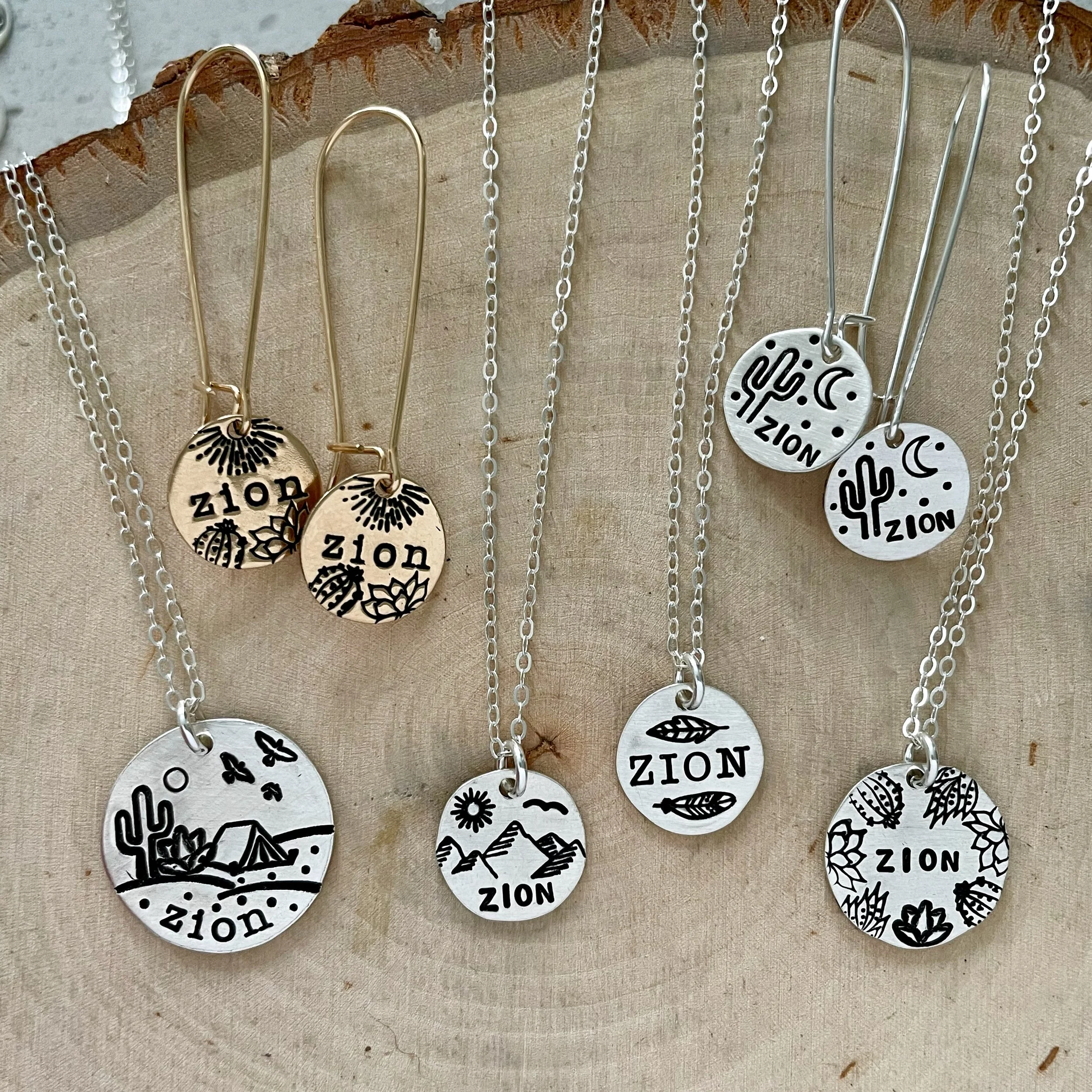 Close-up of silver and gold necklaces hanging on a wooden display, some with round pendants inscribed with "ZION" and decorative designs, and others with various engraved patterns, against a beige fabric background.