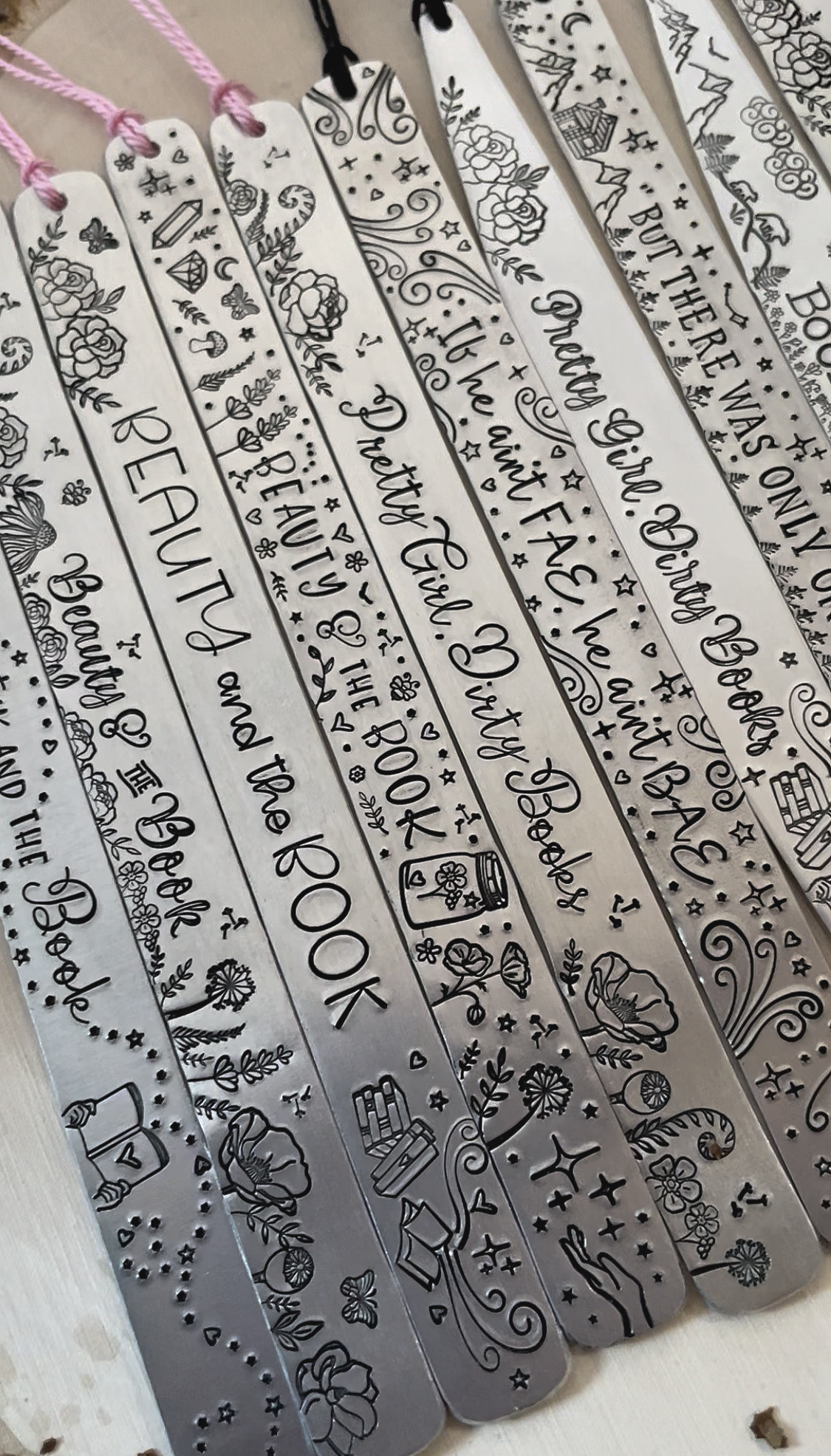 Set of engraved metal bookmarks with various inspirational quotes and decorative illustrations, hanging from pink and black strings.