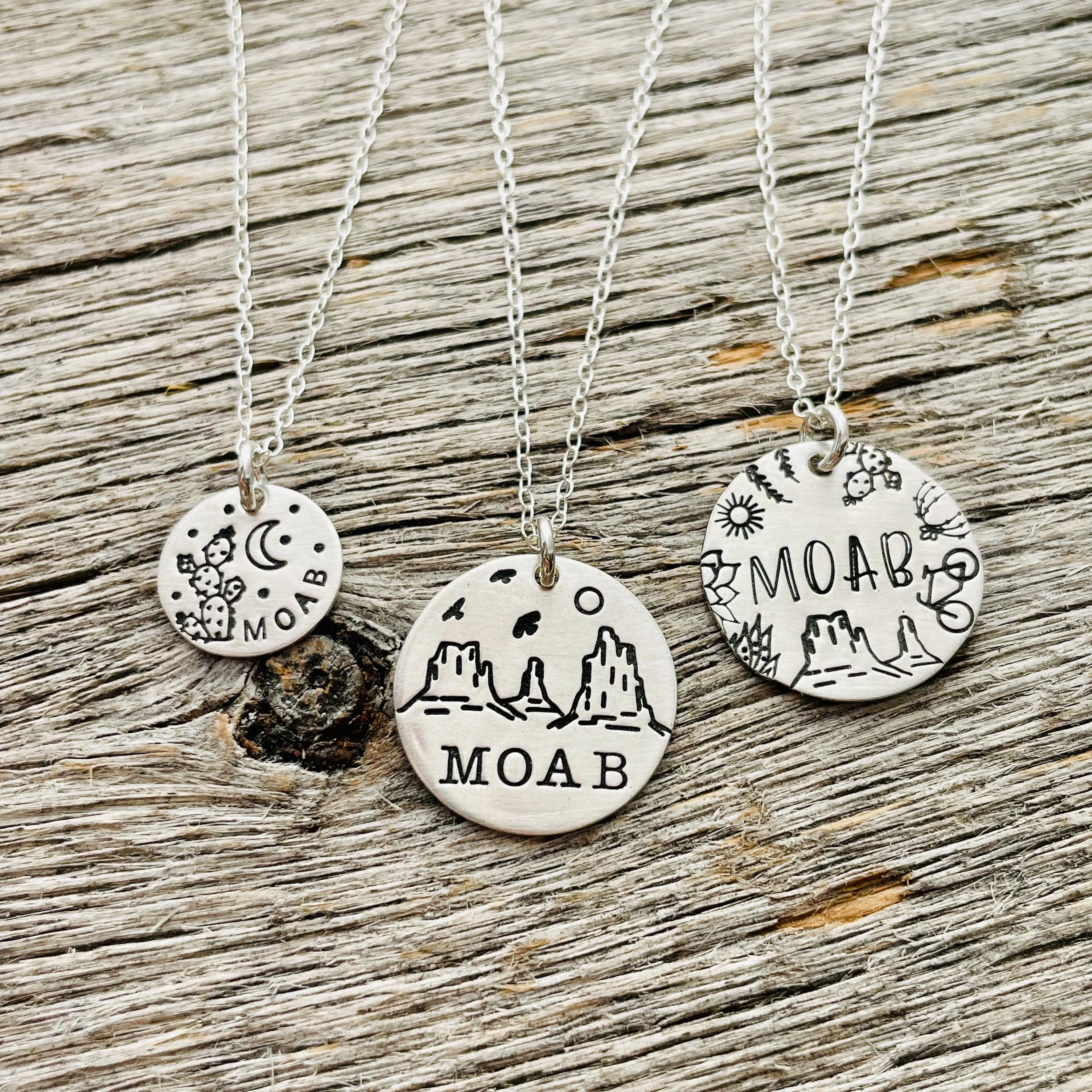 Close-up of white necklaces with circular pendants featuring moon, star, and desert mountain designs, displayed on weathered wooden surface.