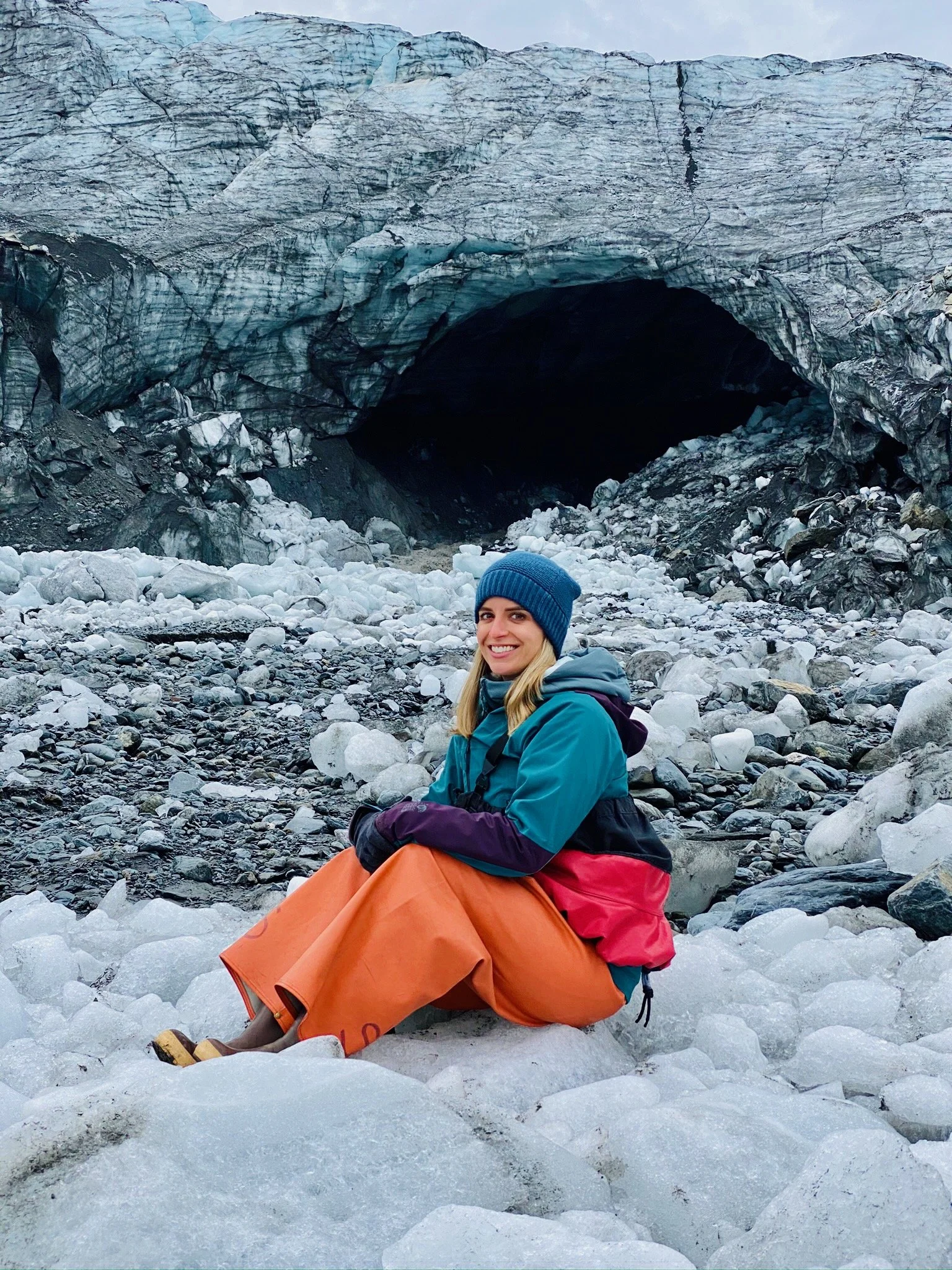 A smiling woman dressed warmly in blue and purple, sitting on snow and ice near a large glacier with a dark ice cave entrance in the background.
