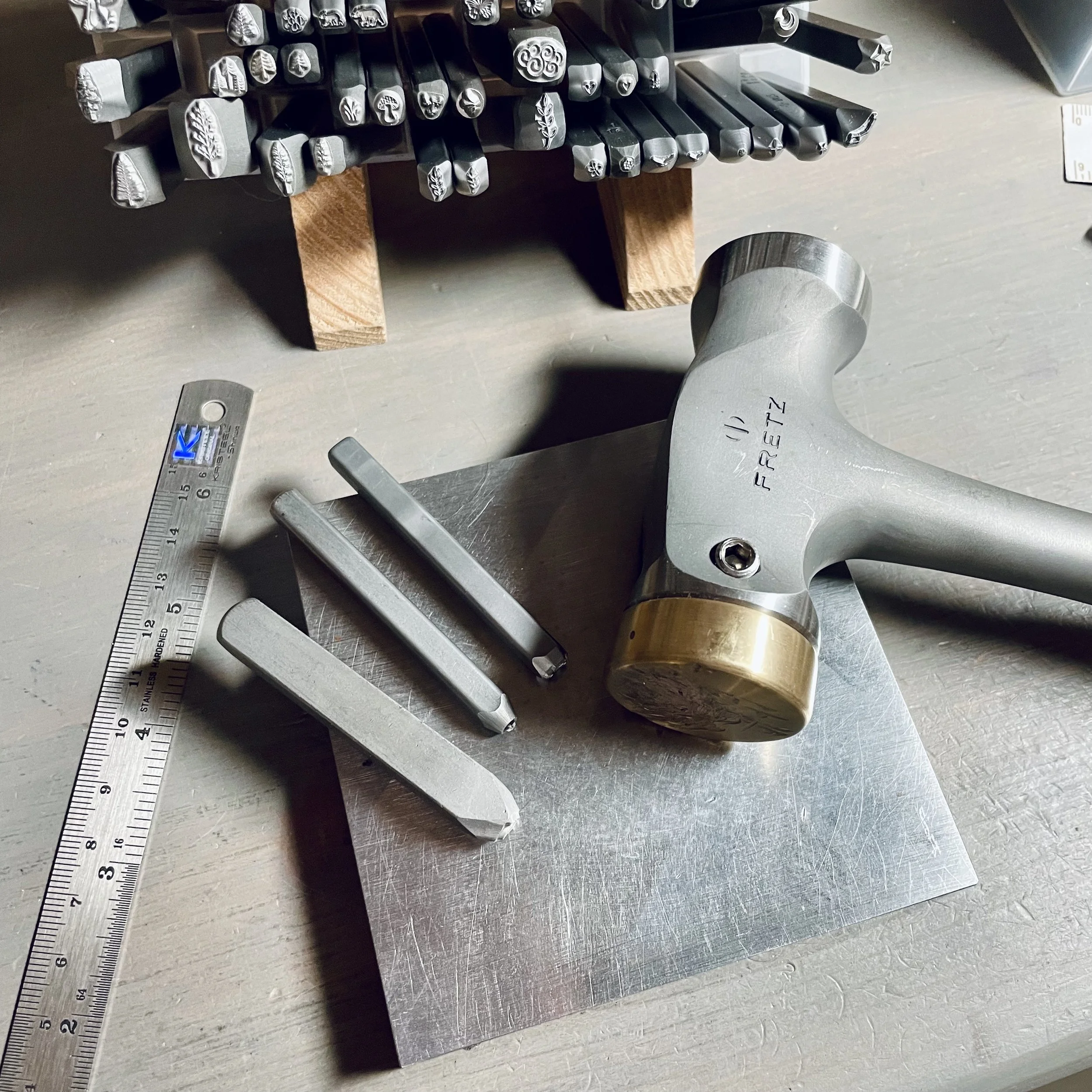 Various metal stamps with engraved symbols and designs, placed on a metal surface, along with a ruler, a hammer, and other metal tools.