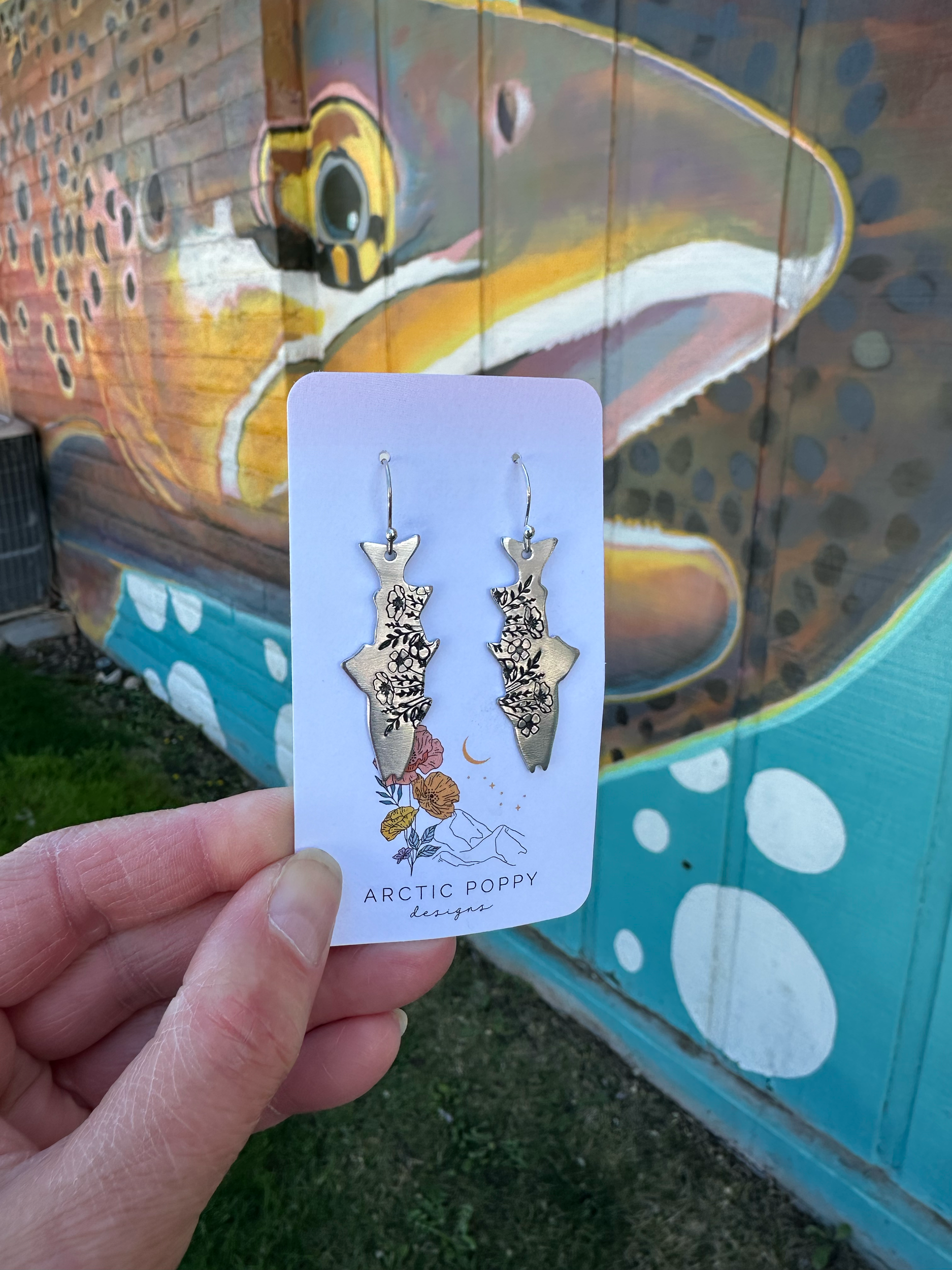 Hand holding a jewelry card with floral earrings in front of a colorful mural of a yellow submarine.