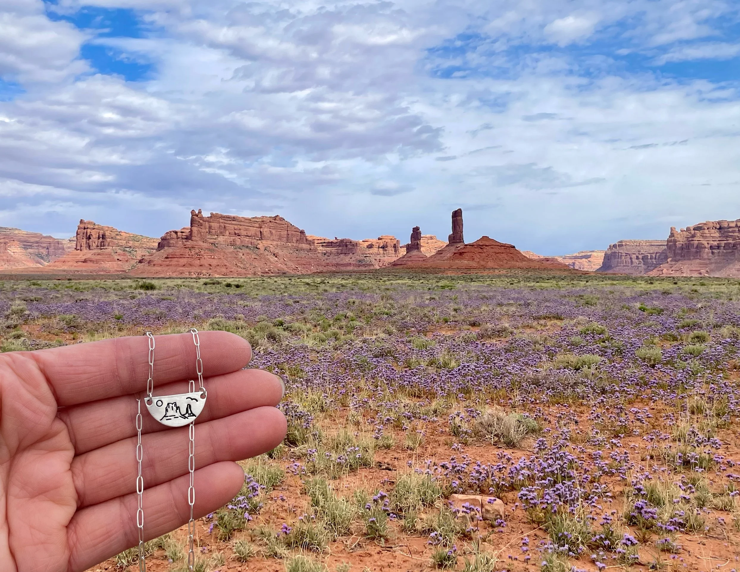 A hand holding a necklace with a pendant that has a mountain and desert scene, set against a desert landscape with red rock formations and purple wildflowers under a partly cloudy sky.