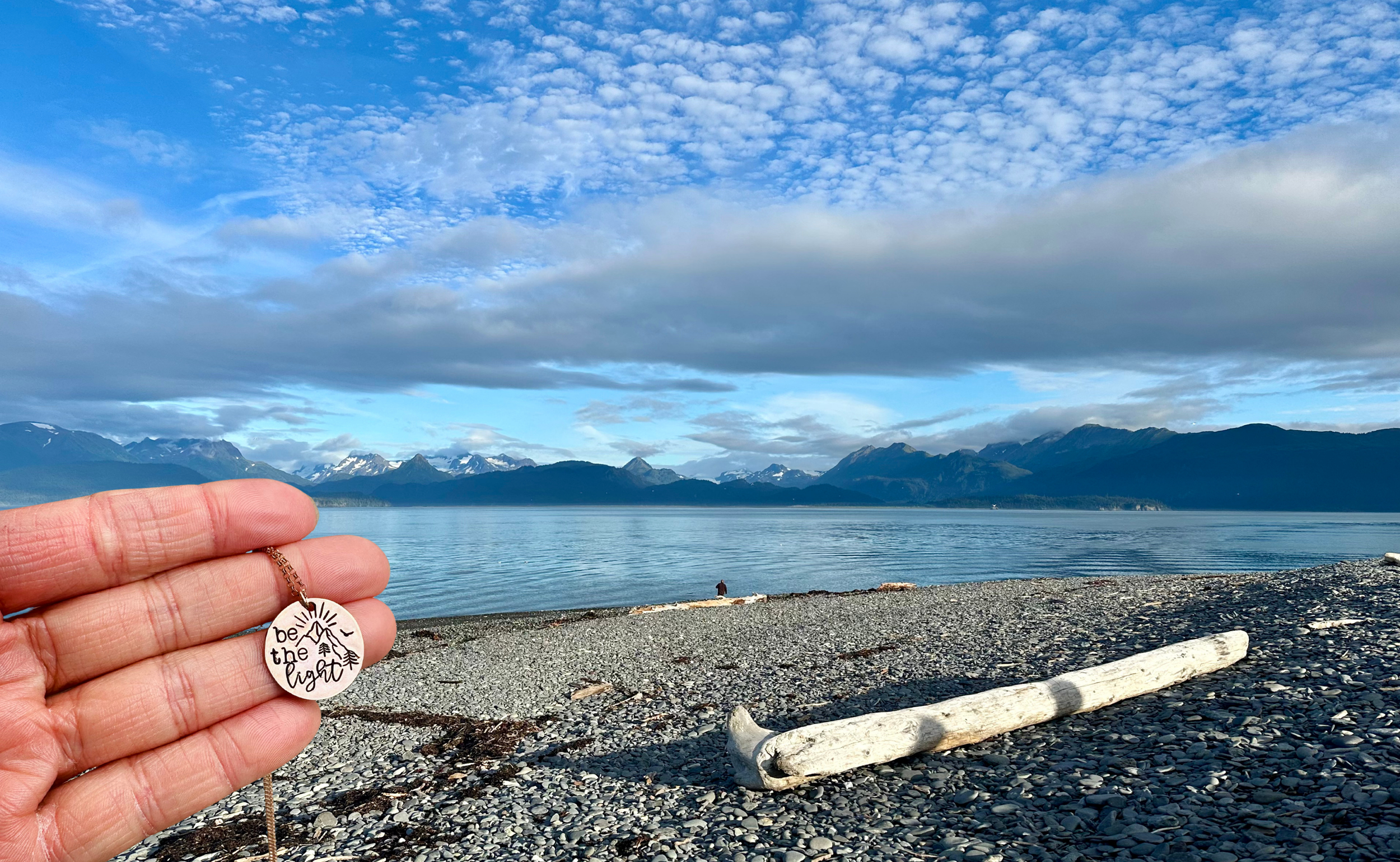 Person holding a necklace with a round pendant that says "believe the light" in front of a scenic view of a lake, mountains, and a blue sky with clouds.