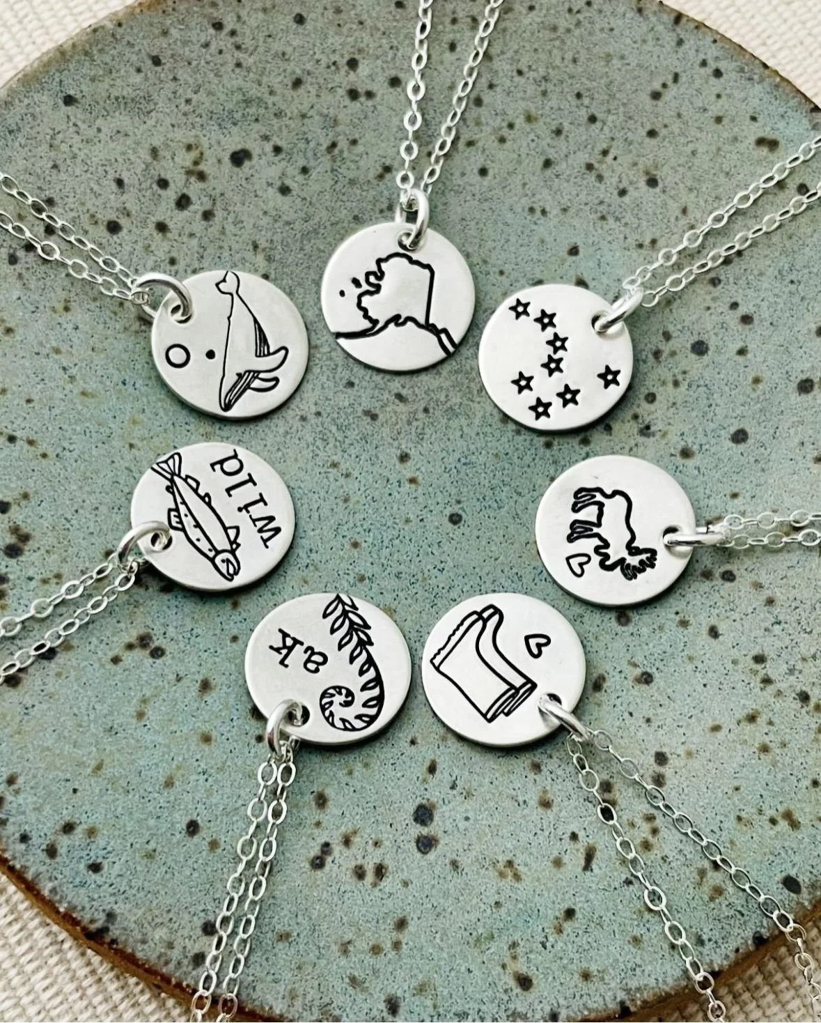Set of seven silver necklaces with circular pendants featuring black line art and text, arranged on a textured, speckled surface.