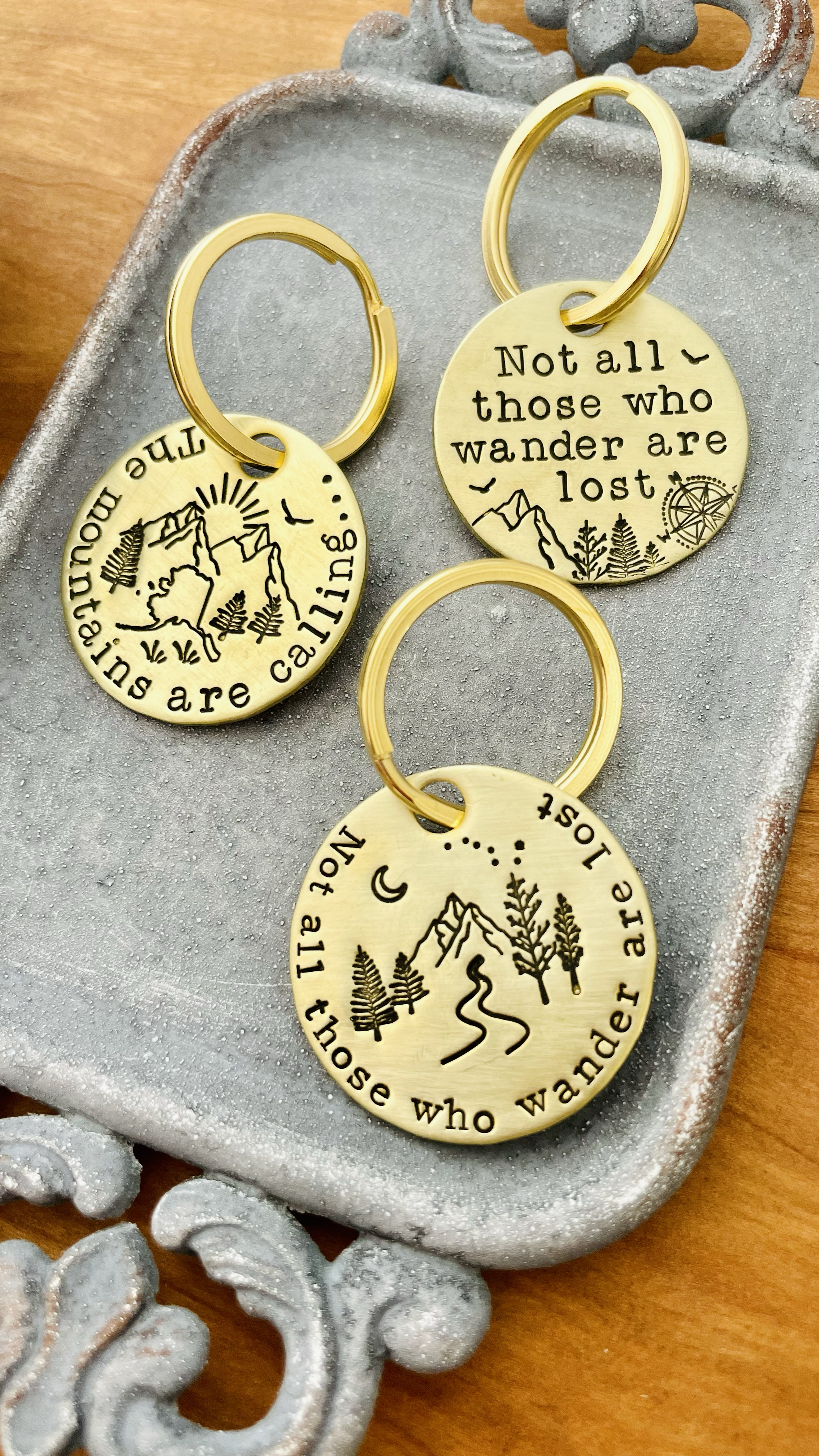 Gold-colored keychains with engraved mountain, pine trees, and quote about wandering on a textured surface.
