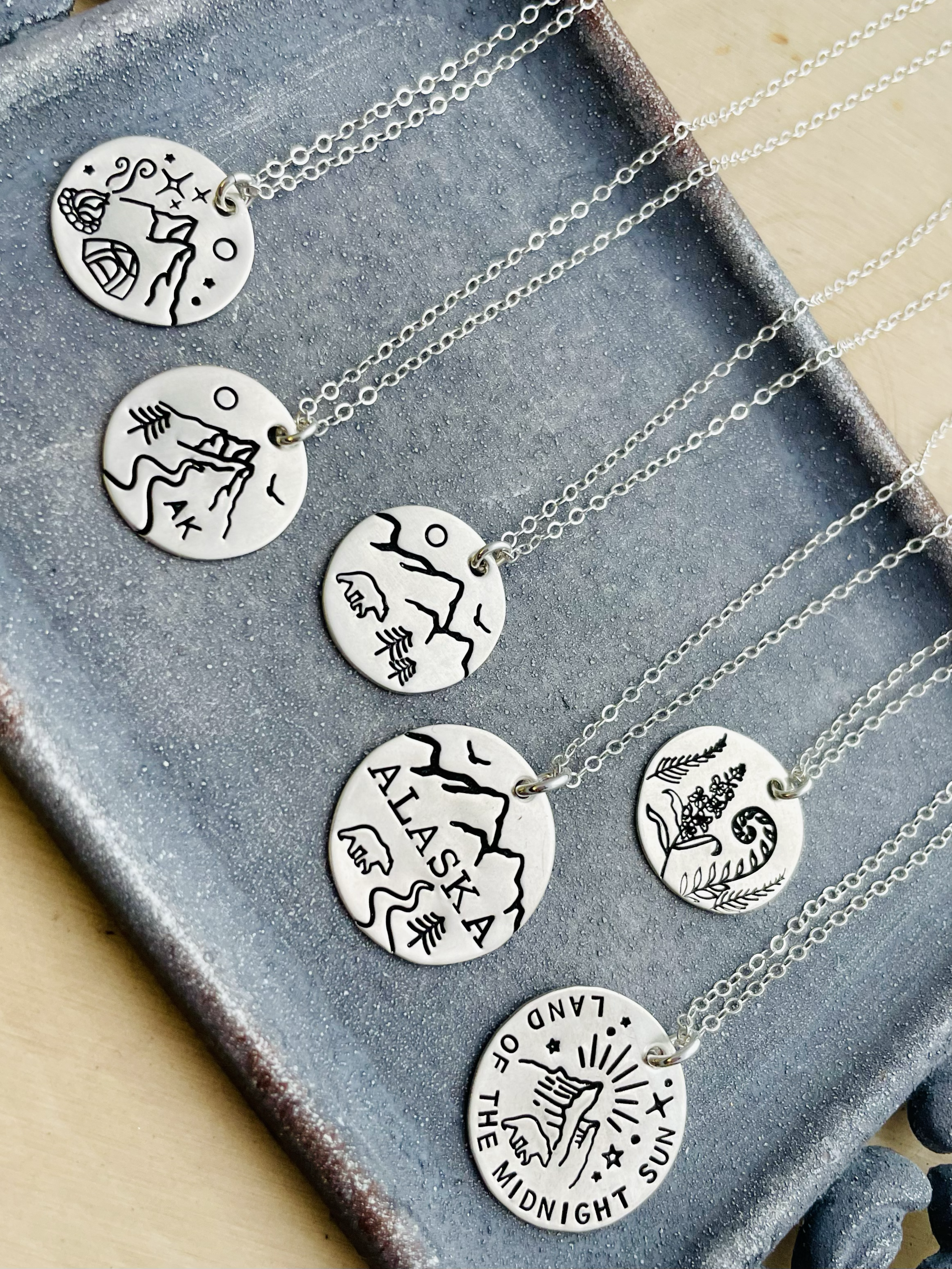 Silver necklaces with circular pendants featuring outdoor and camping-themed illustrations and text, displayed on a textured surface.