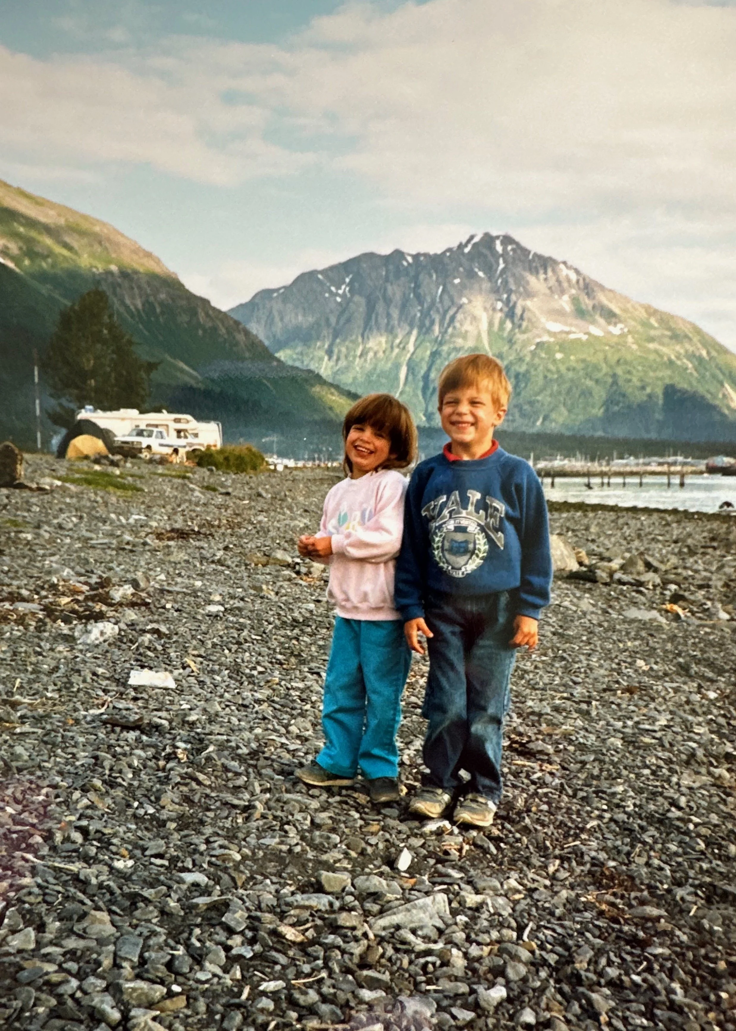 Two children, a girl and a boy, standing on a rocky beach with mountains and a marina in the background, smiling.