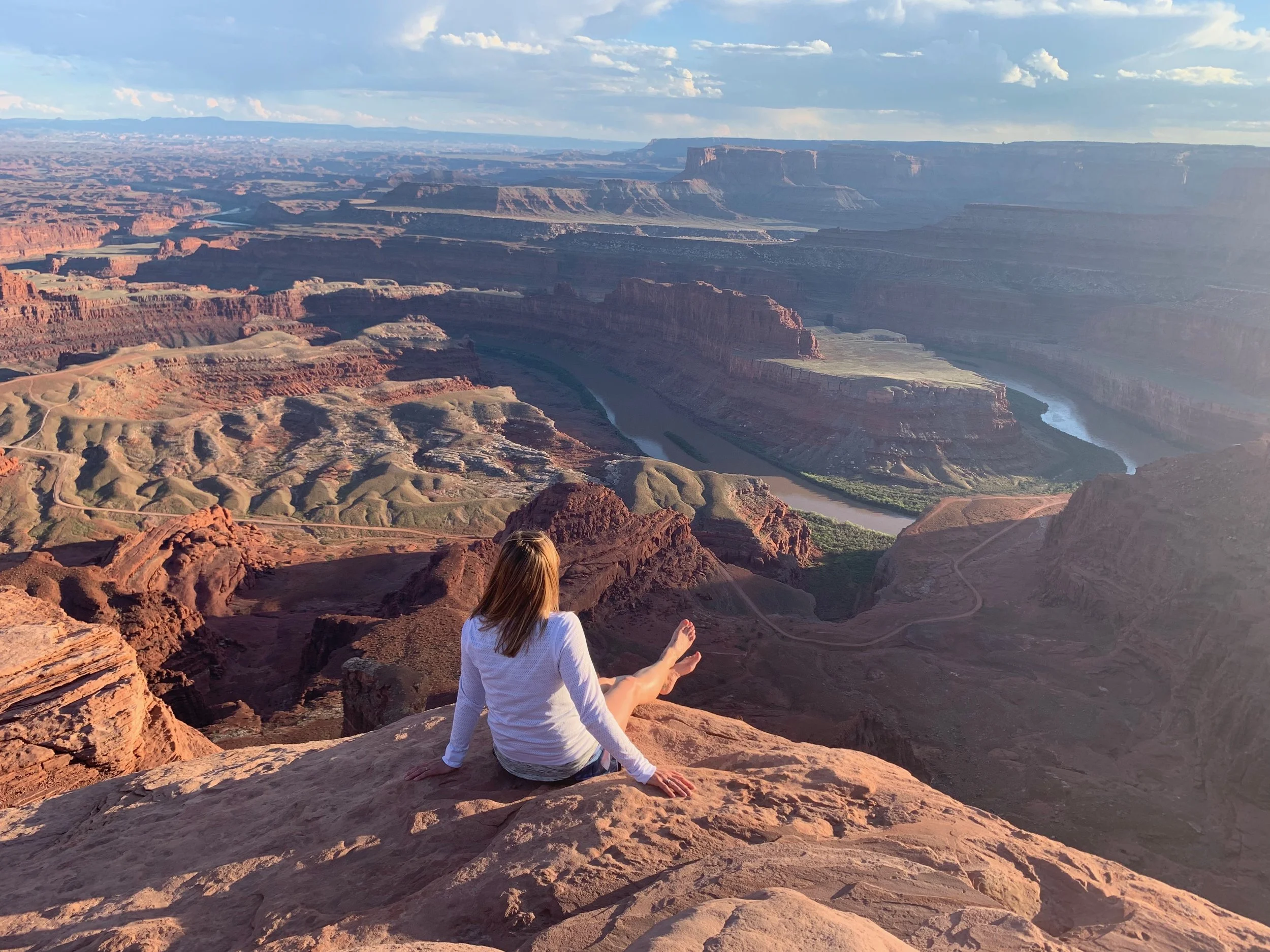 A person in a white shirt sitting on rocks at the Grand Canyon, looking out at the vast canyon and the Colorado River below, with a view of layered red and orange cliffs and distant plateaus under a partly cloudy sky.