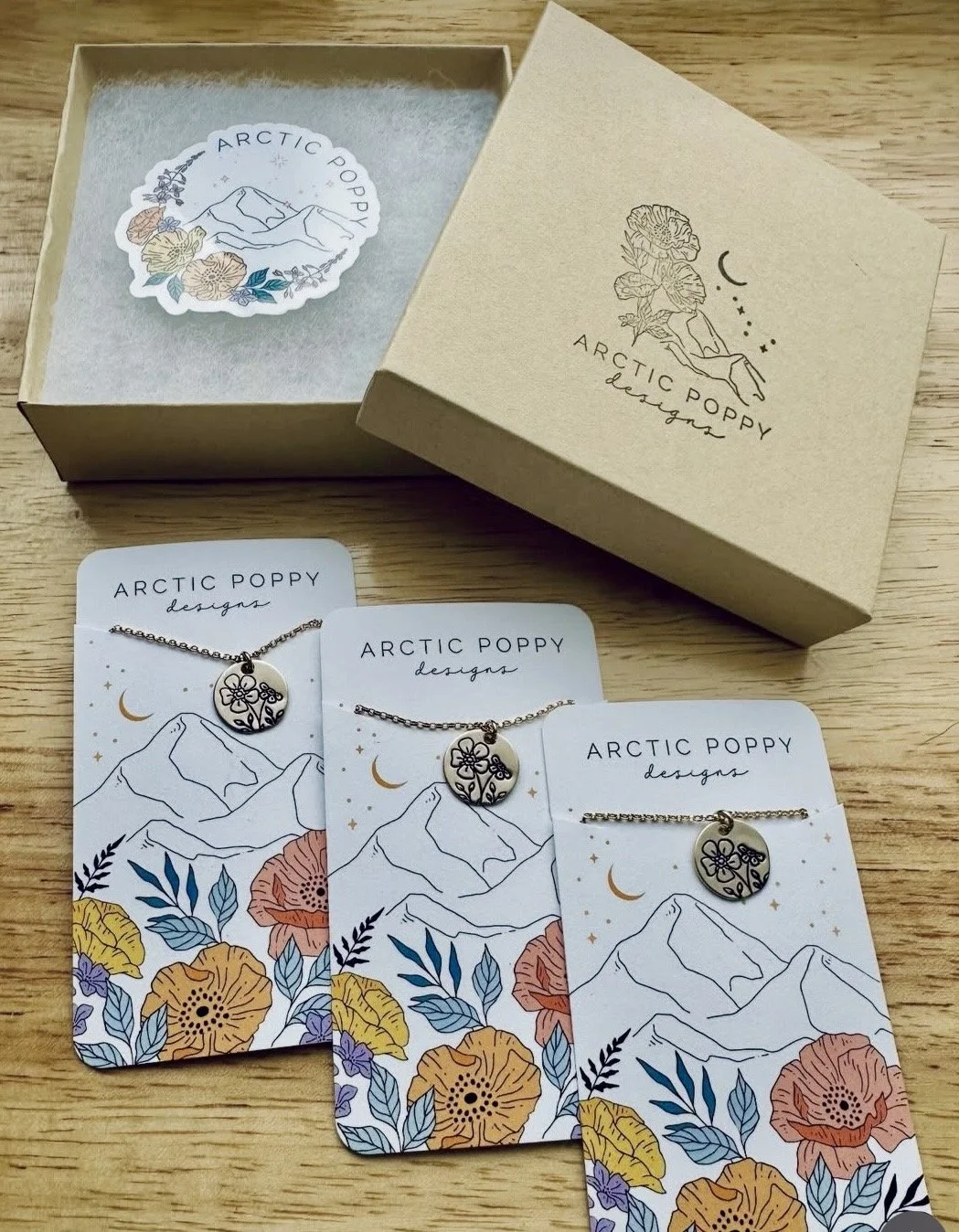Three jewelry necklaces with floral and mountain designs on branded cards, and a gold box with a mountain and nature design for Arctic Poppy Designs, on a wooden surface.