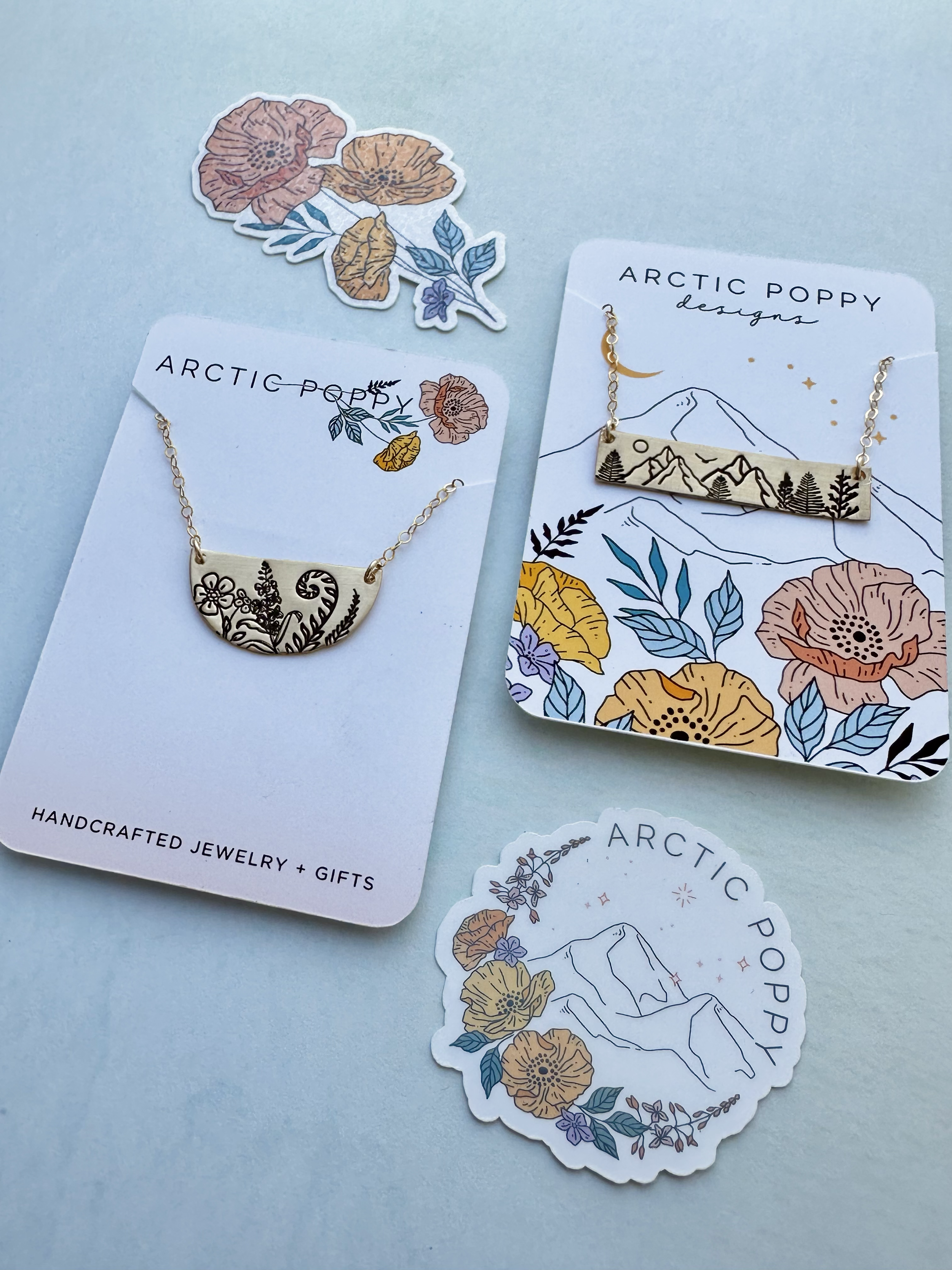 A collection of handmade jewelry and stickers with floral and mountain designs on a light blue background. The jewelry includes necklaces with mountain and floral motifs. The packaging features branding from Arctic Poppy Designs with matching floral 