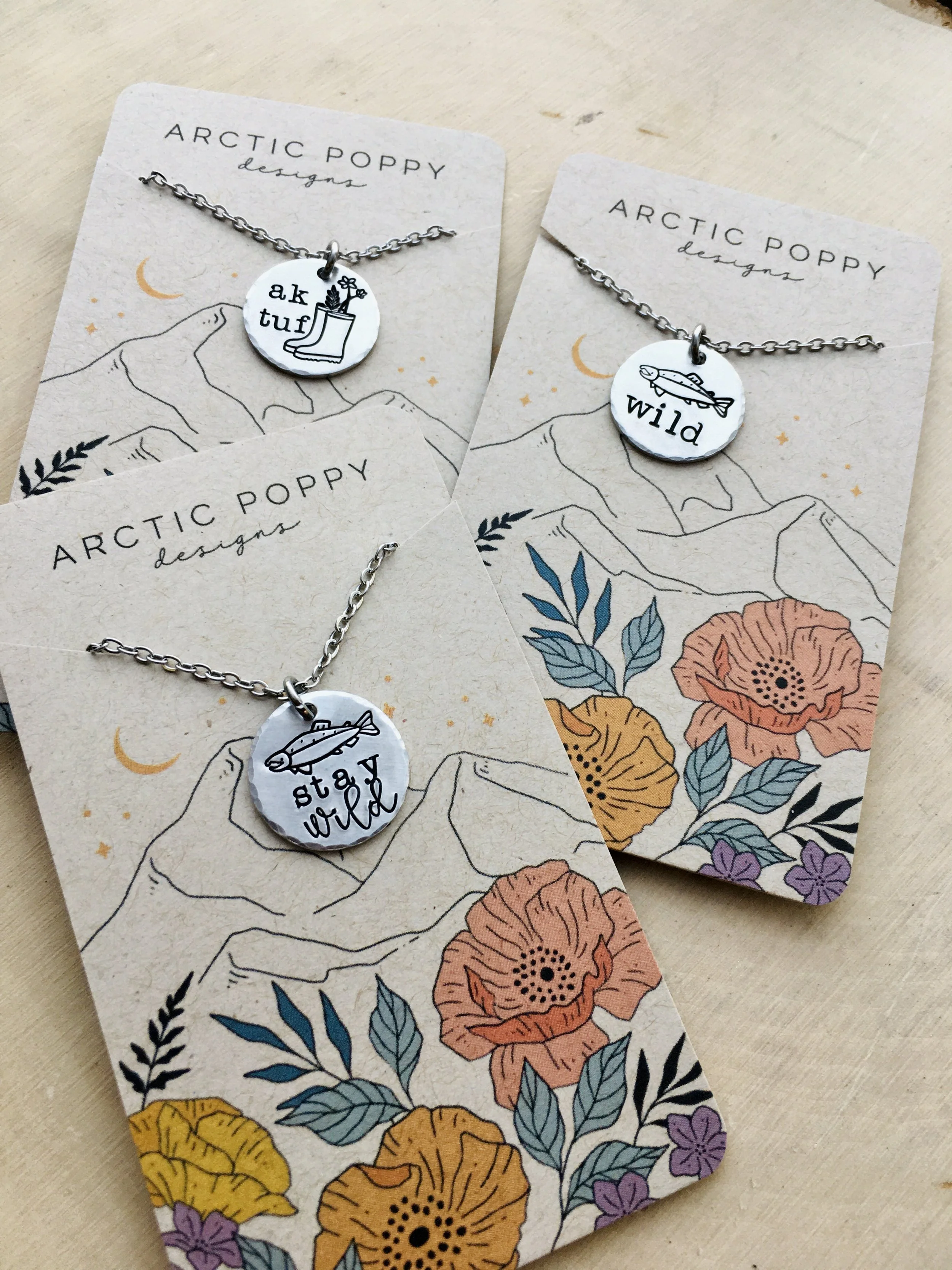 Two jewelry cards with silver necklaces, each featuring a round charm with black illustrations and text, placed on a floral and mountain illustrated background.