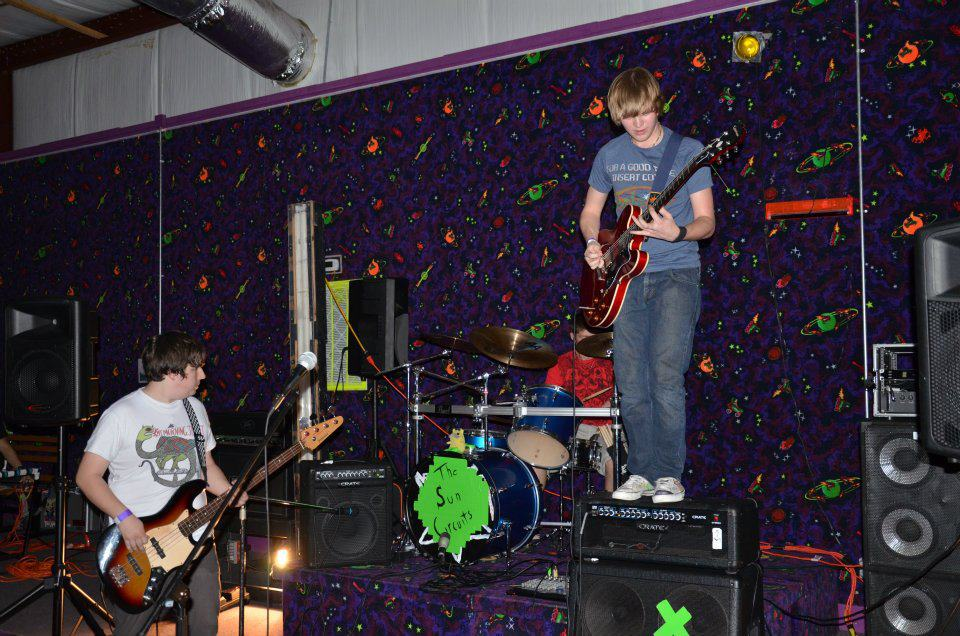 Two young boys playing guitars on a stage with a drum set behind them, in a room with space-themed wallpaper. The boy on the right is standing on an amplifier, playing an electric guitar, while the boy on the left is playing a bass guitar. There are large speakers and other musical equipment on stage.