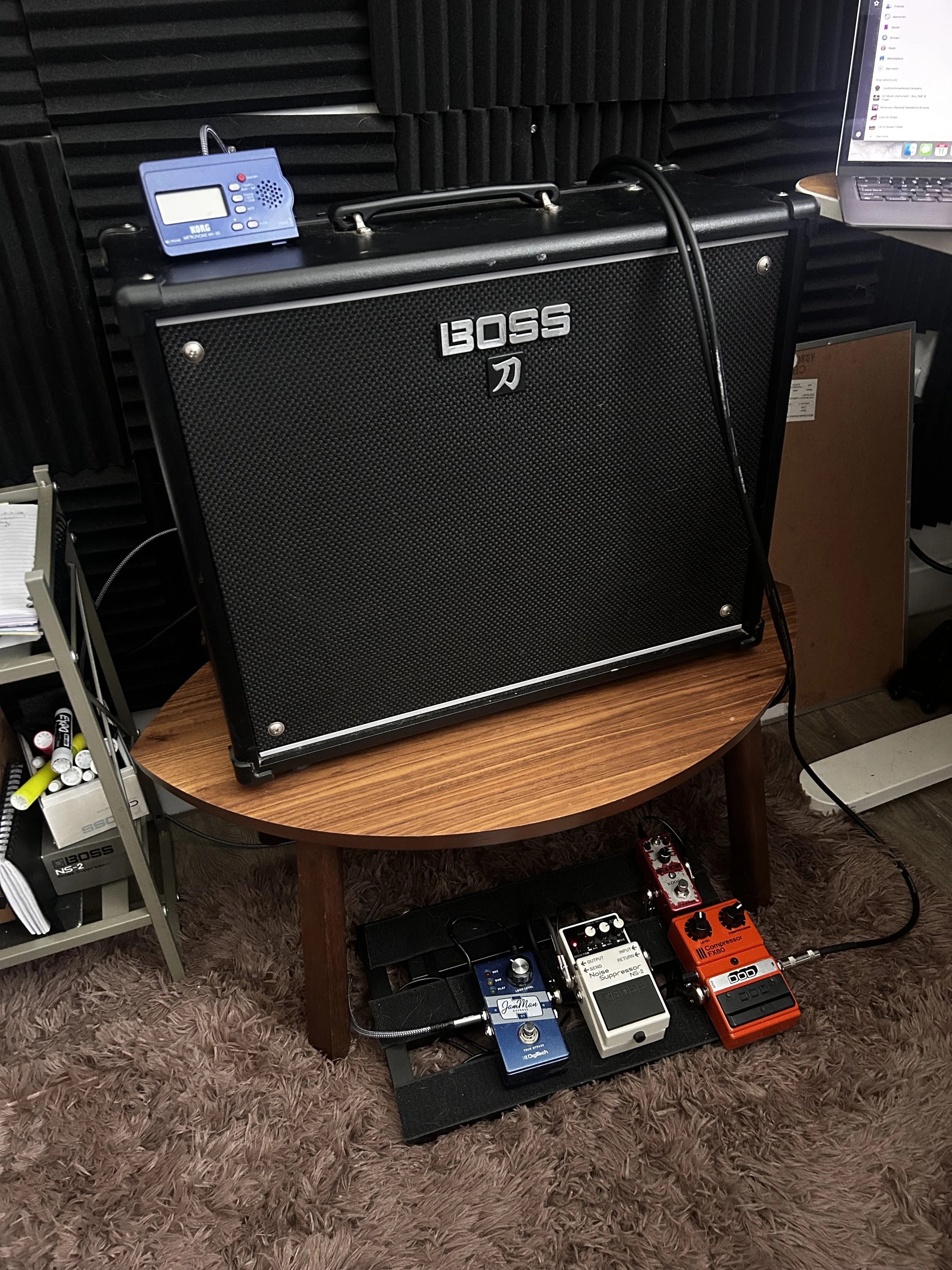 Photo of a guitar amplifier with various effects pedals and a laptop on a table, music gear room with acoustic foam panels.