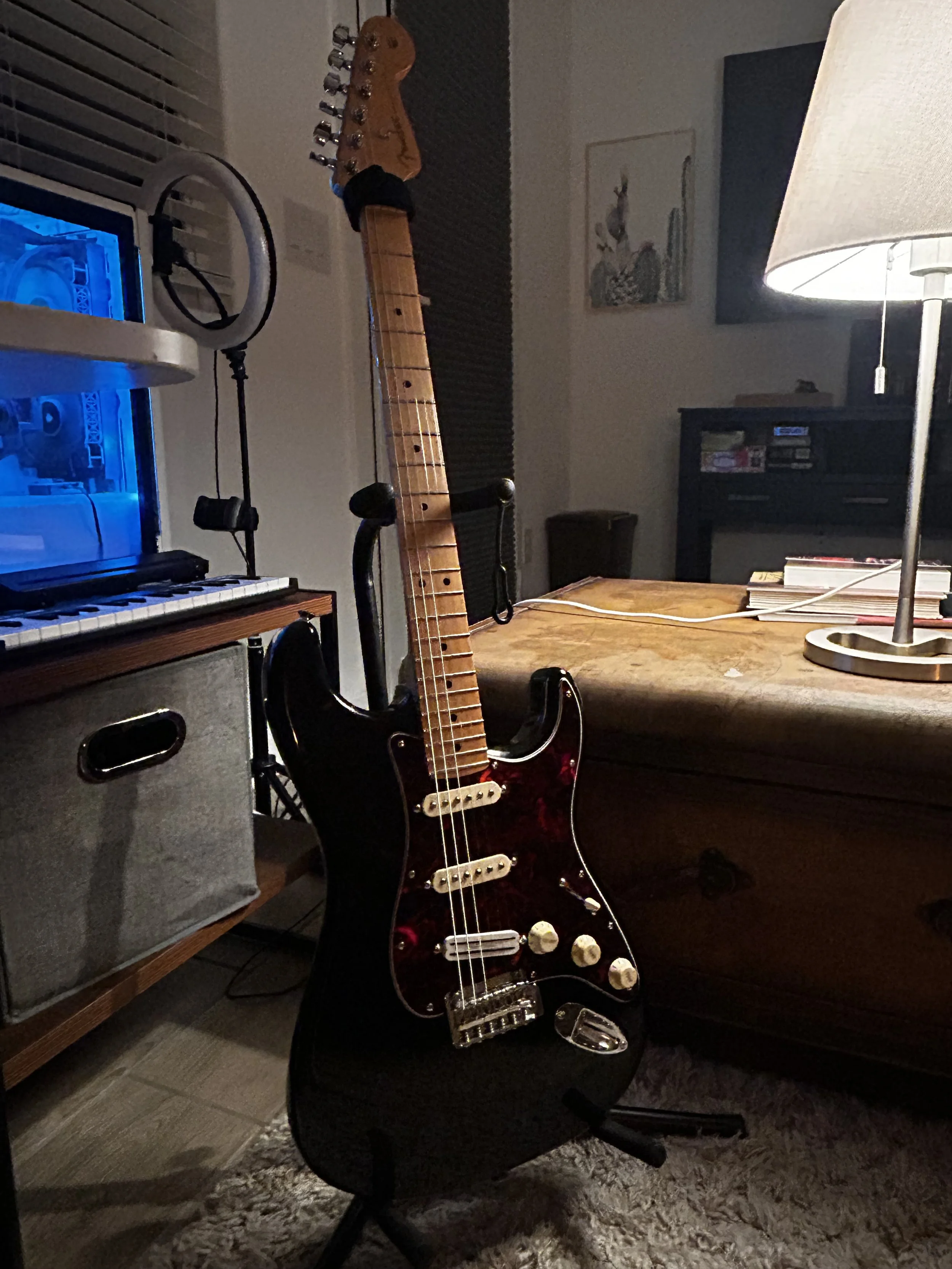 An electric guitar with a black body, white pickups, and four control knobs is leaning against a black chair in a room with a wooden desk, a table lamp, and a framed artwork of a rabbit on the wall.