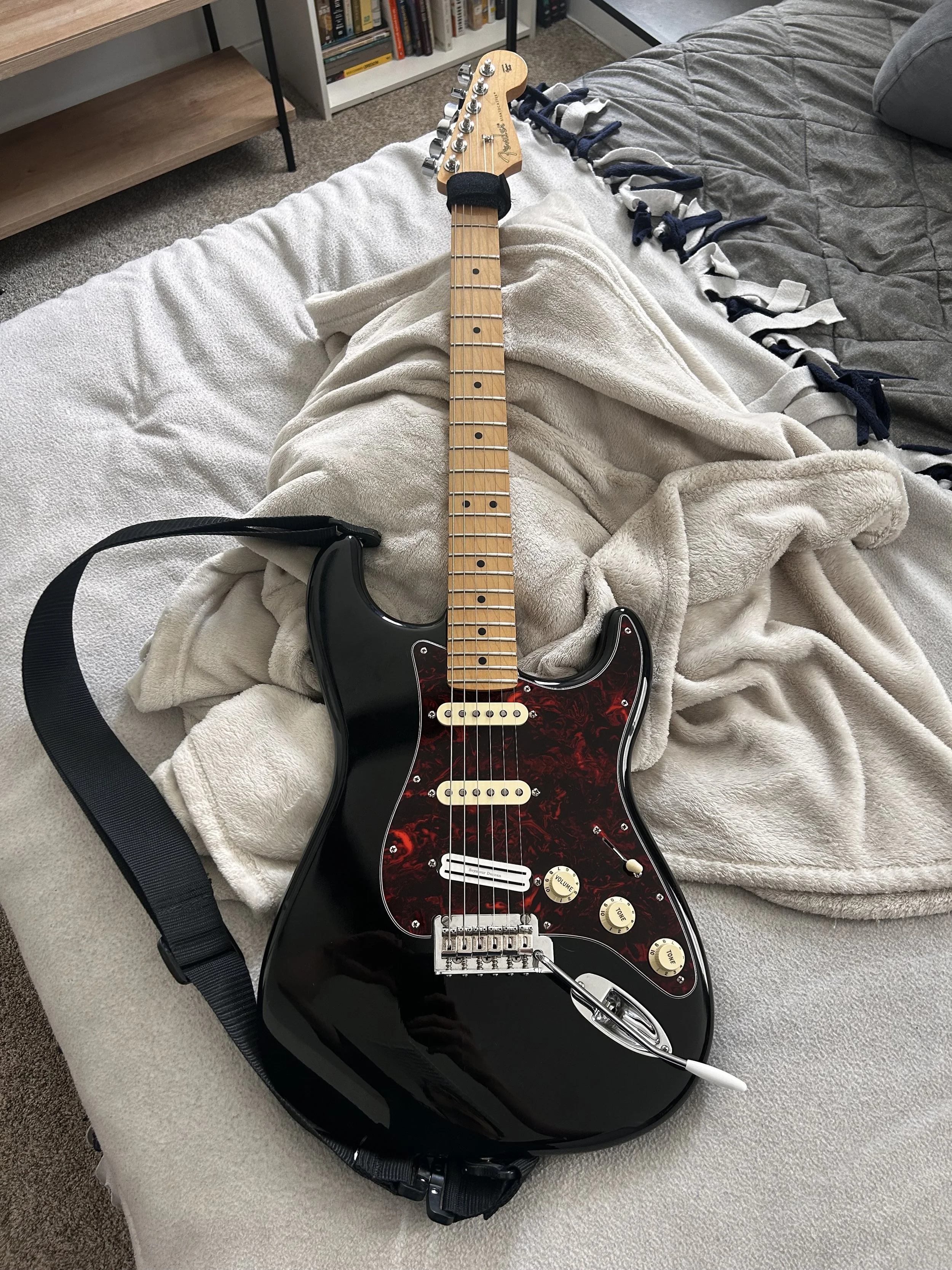 Electric guitar with a black body, red tortoiseshell pickguard, and maple neck, resting on a bed with a beige blanket.