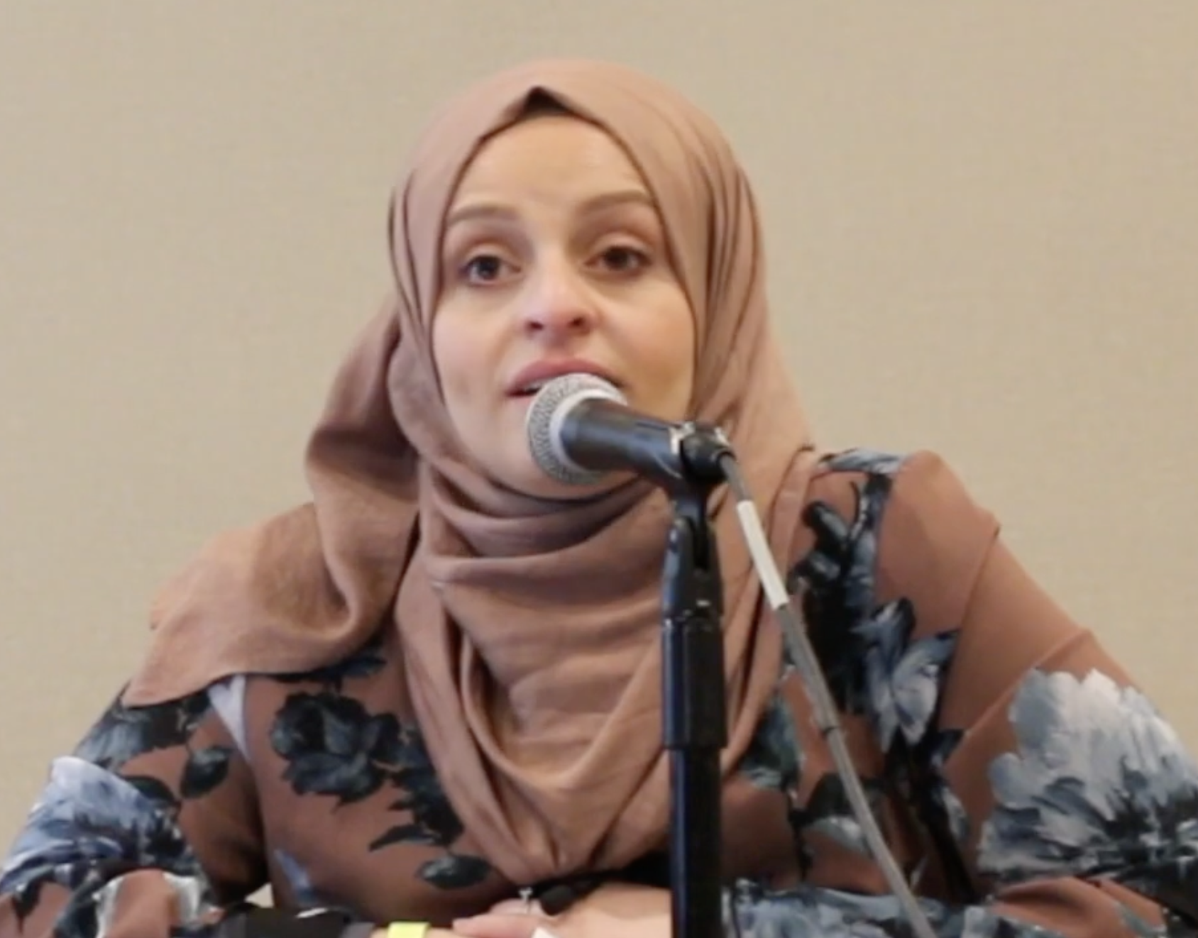 A woman wearing a beige hijab and floral blouse speaking into a microphone at a panel or event.