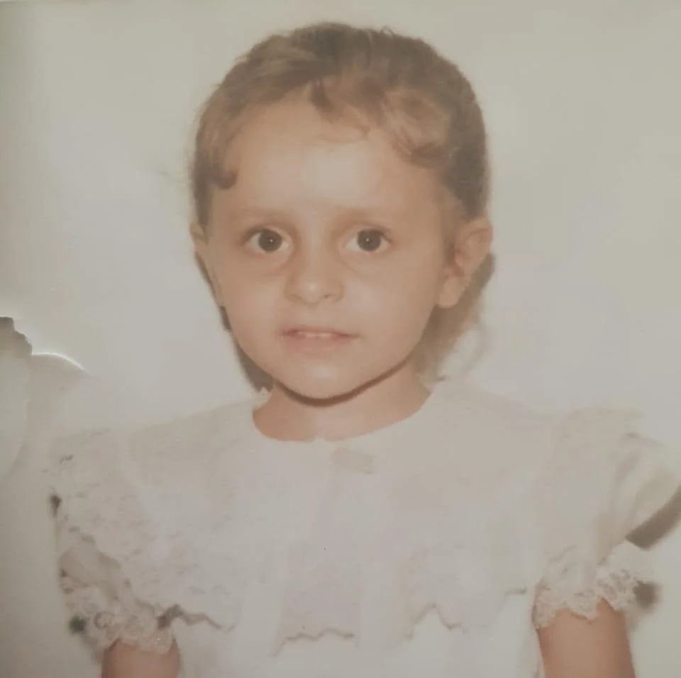 A young girl with short brown hair and large eyes, wearing a white dress with lace details, standing against a plain background.