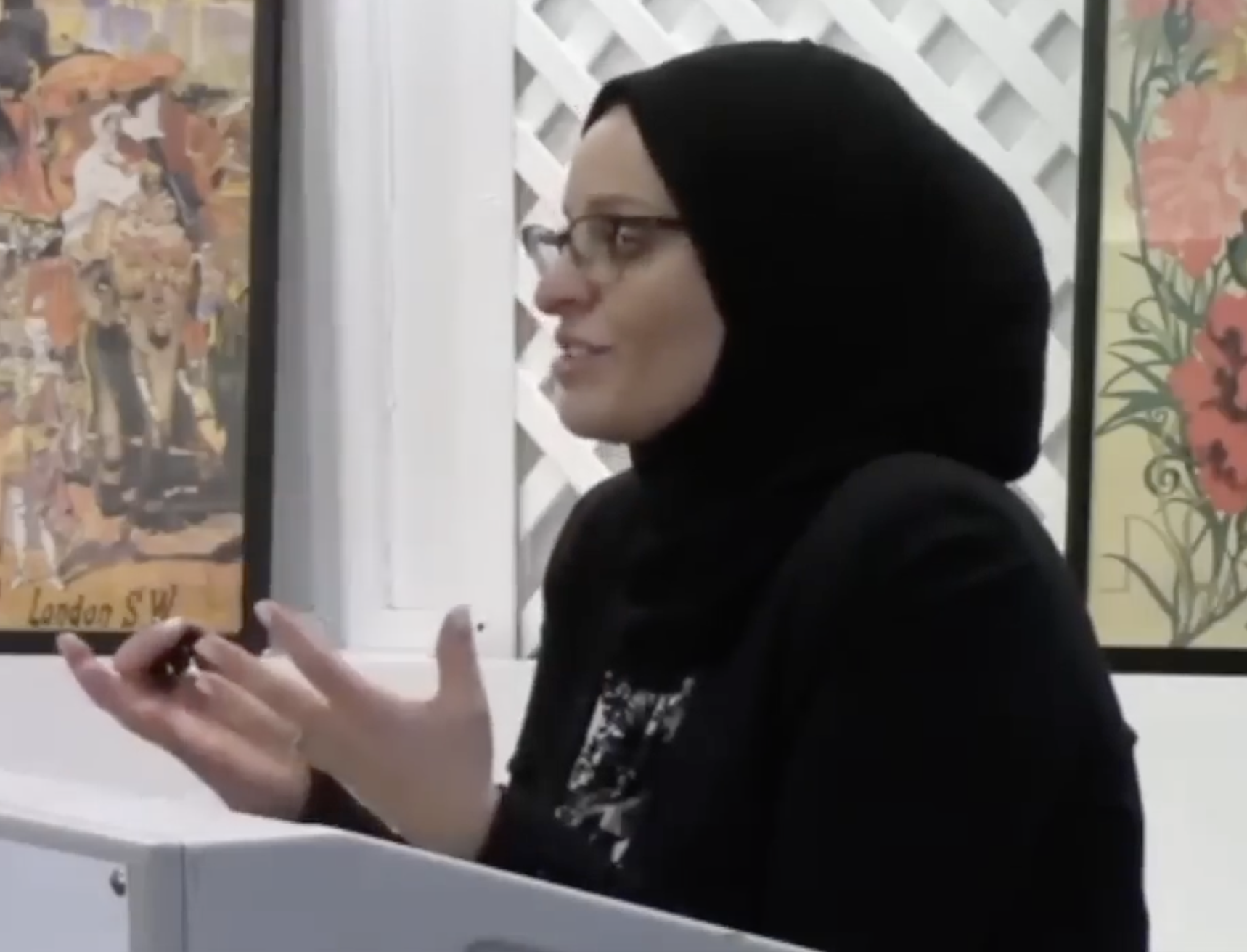 A woman with glasses and a black hijab is standing indoors, gesturing with her hands, with framed artwork hanging on white walls behind her.