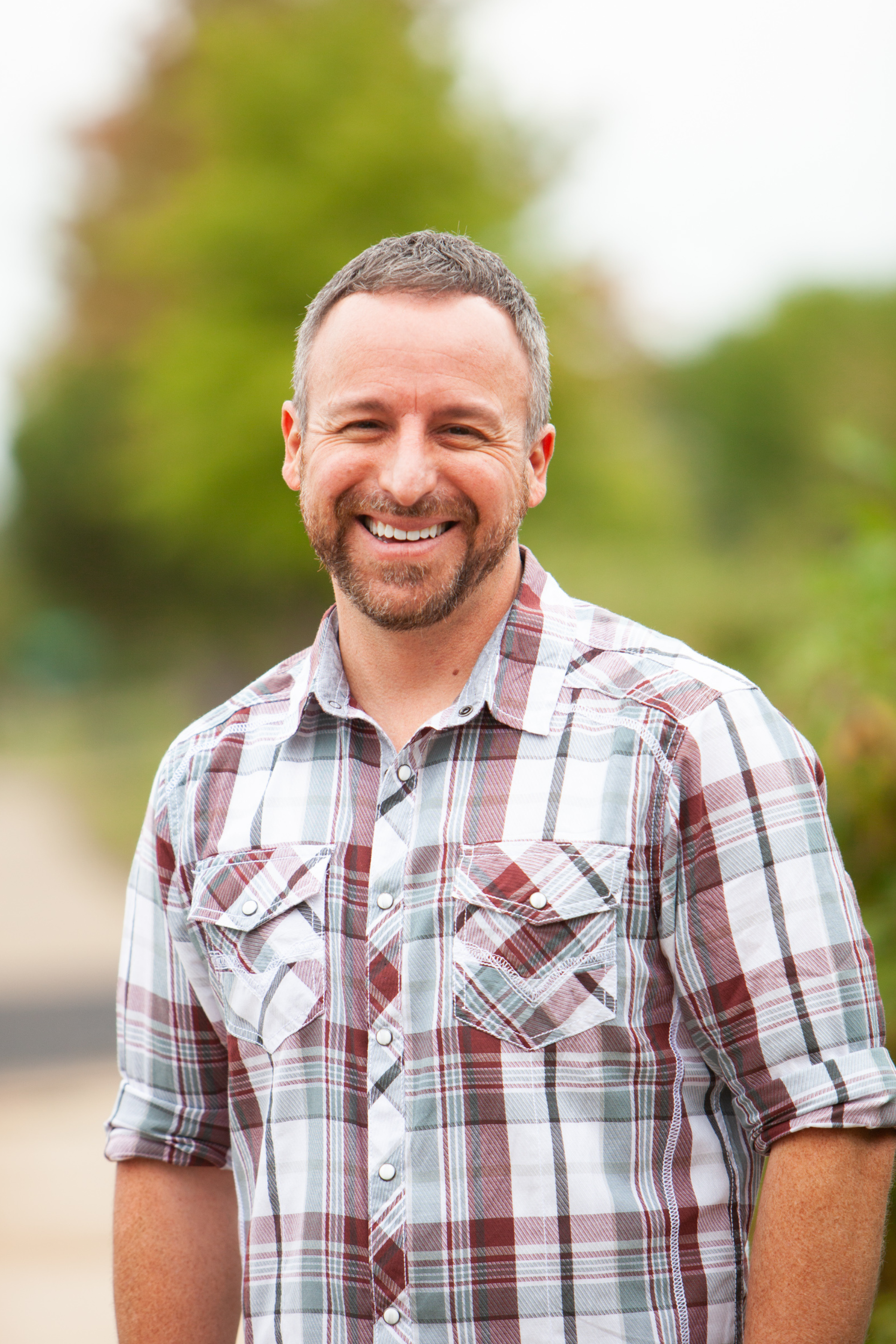 A smiling man with short hair and a beard wearing a plaid shirt outdoors with trees in the background.