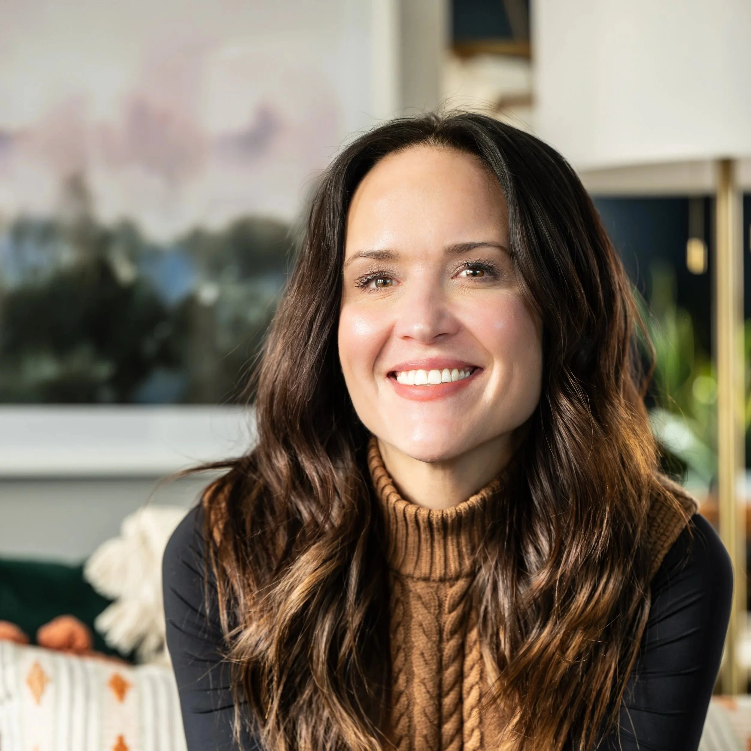 Close-up of a woman with long brown hair smiling, wearing a brown turtleneck sweater and a black jacket, in an indoor setting with shelves and blurred decorative items in the background.
