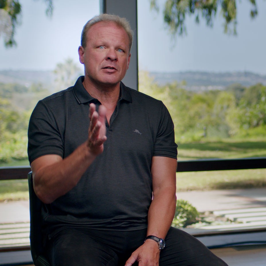 A man with light-colored hair and fair skin sitting indoors near a large window, outdoors view of green trees and landscape, wearing a black short-sleeve polo shirt and a watch.