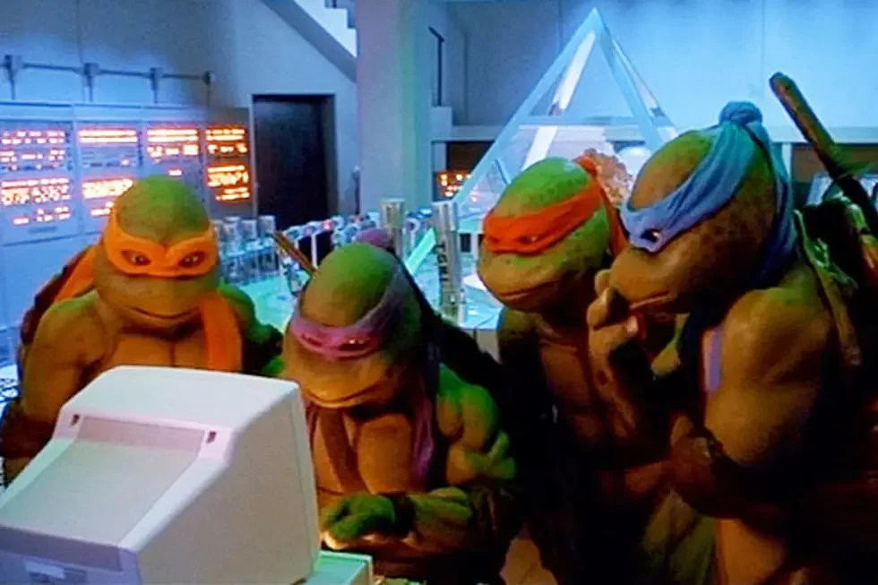 Illustration of four movie-style Ninja Turtles gathered around a computer in a blue-lit room with a glass pyramid in the background
