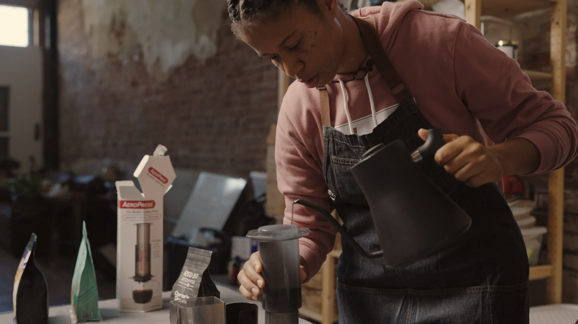 Person pouring water from a black kettle into a cup, with coffee-making supplies on a table, in a workshop setting with exposed brick walls.
