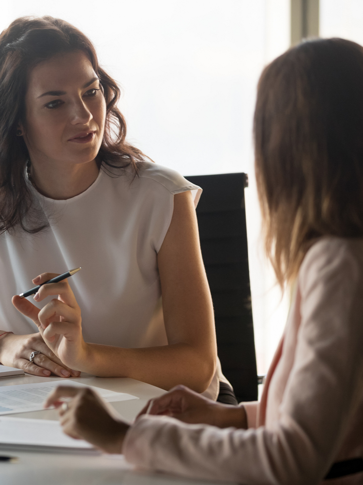 Two women having a discussion at a desk, one holding a pen, in an office setting.
