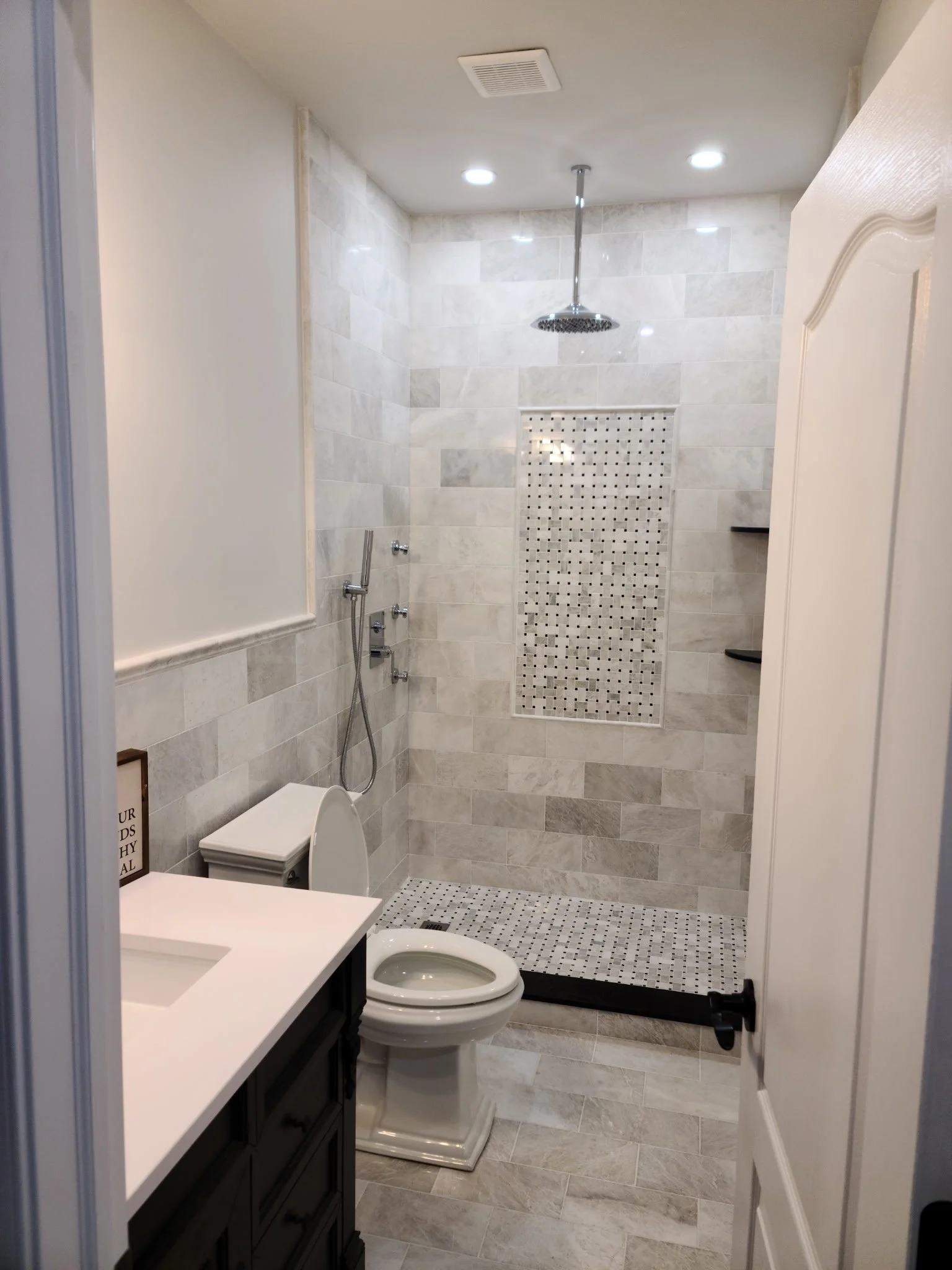 A small bathroom with a modern shower area, a toilet, and a vanity with a sink. The shower has a rainfall showerhead and a niche with small mosaic tiles. The walls and floor are tiled in neutral tones.