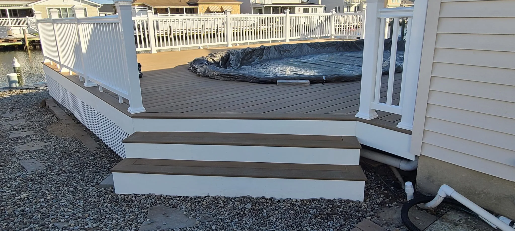 A newly built wooden deck with stairs leading down to the ground, surrounded by a white railing, attached to a house near a body of water with neighboring houses in the background.