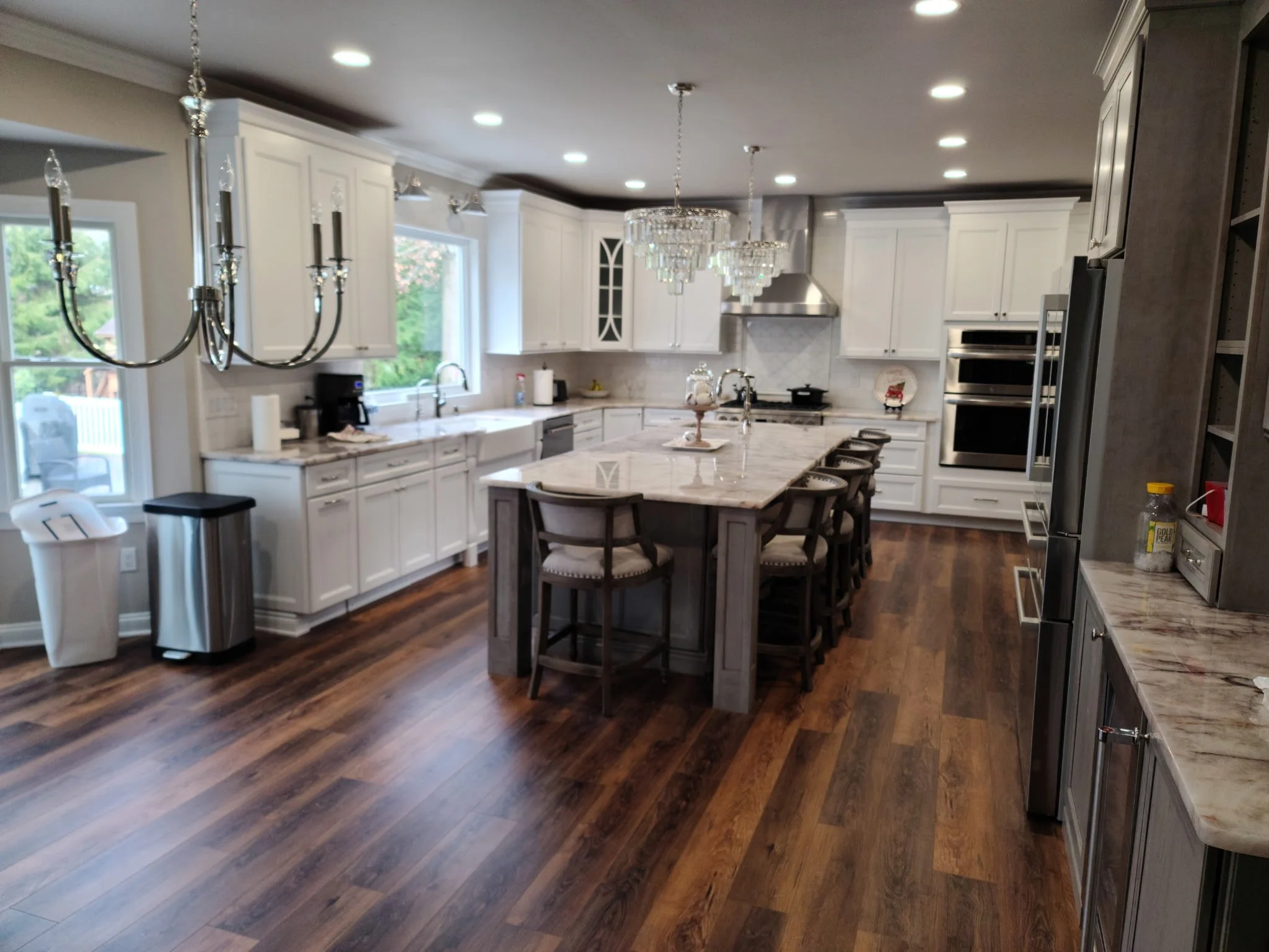 A spacious kitchen with white cabinets, a large central island, hardwood floors, and multiple chandeliers hanging from the ceiling. There is a window over the sink, and kitchen appliances like a double oven and refrigerator are visible.