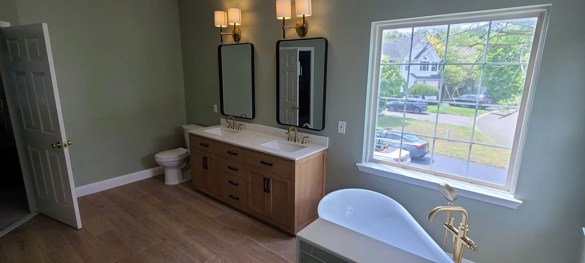 A modern bathroom with a wooden vanity that has two sinks and black hardware, two mirrors above the vanity, a wall-mounted light fixture with multiple lamps, a toilet, a large window showing parked cars and greenery outside, and a white bathtub with a gold faucet in the foreground.