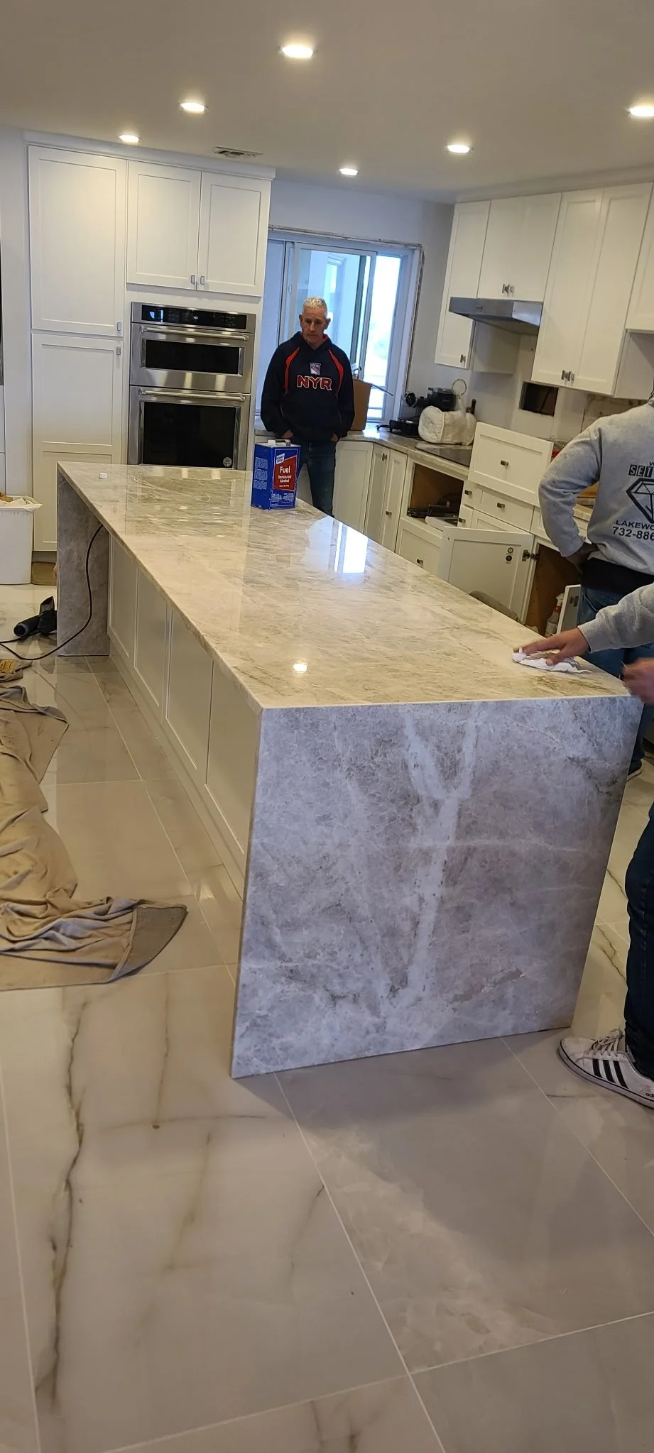 A spacious kitchen with a large marble island, white cabinets, and stainless steel appliances. Two people are present, one in a NYR hoodie and the other in a gray hoodie, working on the island as part of a renovation.