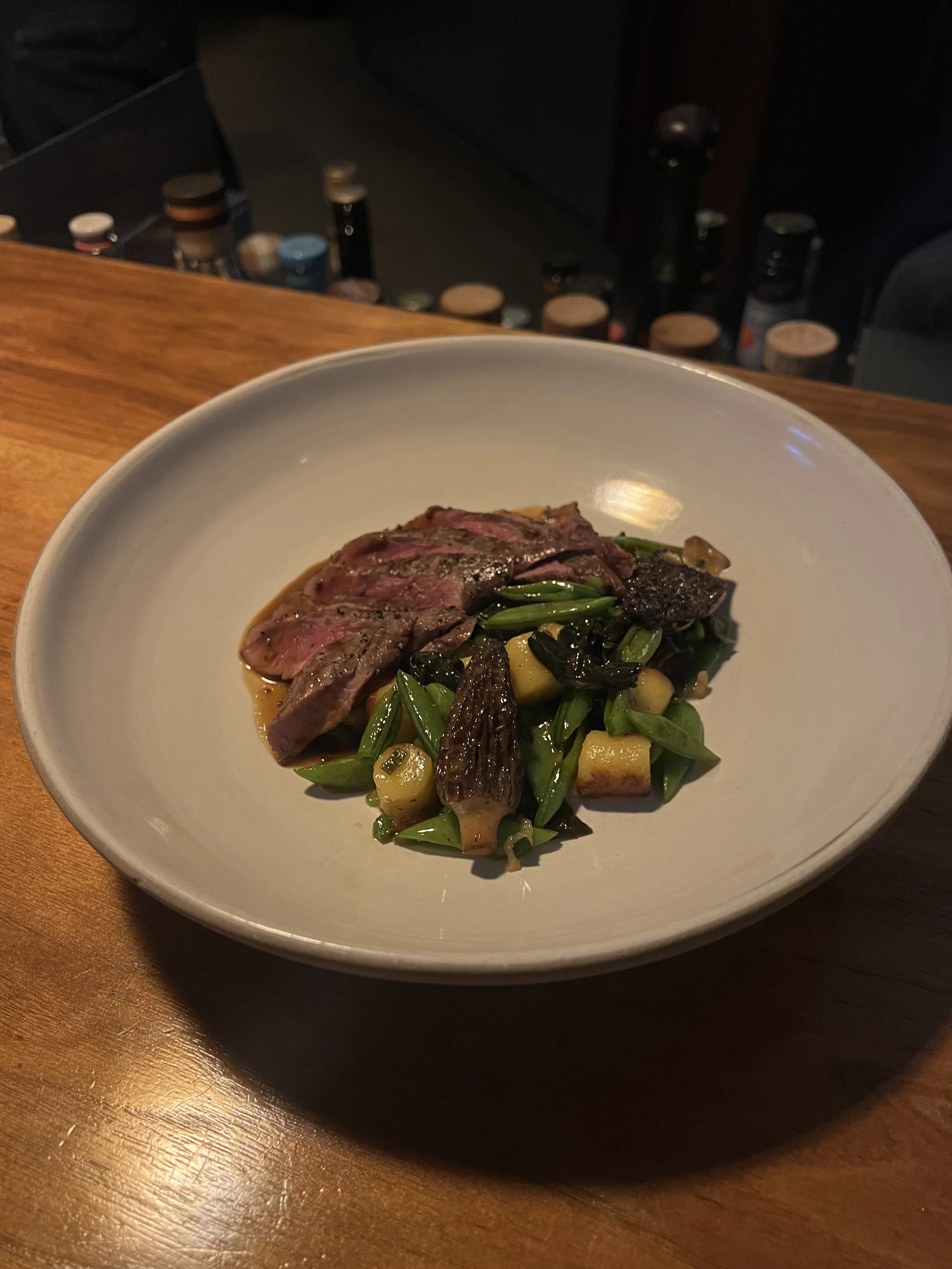 A white bowl with roasted wild boar, spring vegetables, gnocchi romana, and mustard jus on a wooden table.