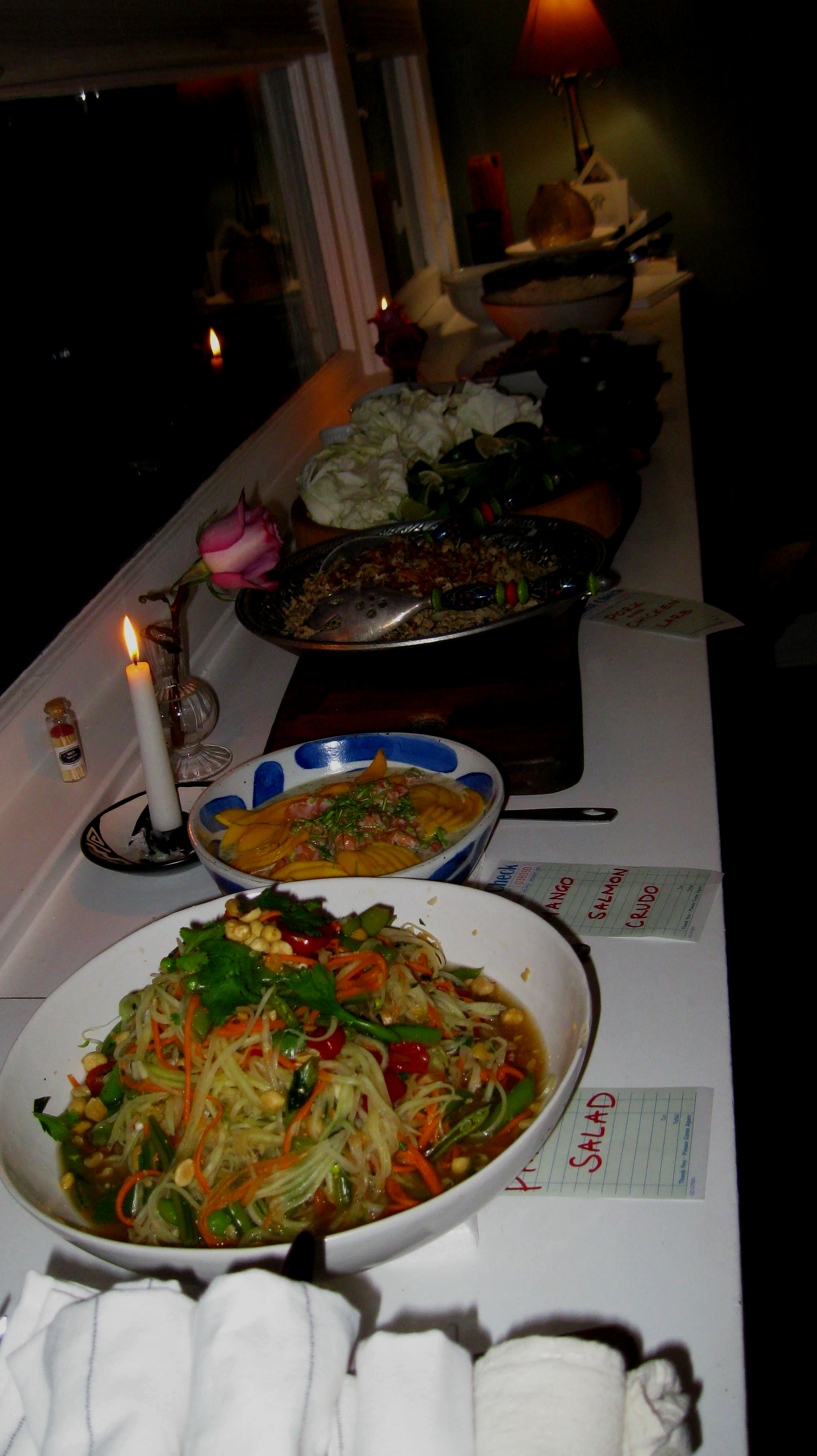 A long buffet table with Thai dishes such as chicken larb, papaya salad, curries, and fresh vegetables. There are candles and flowers as decorations.