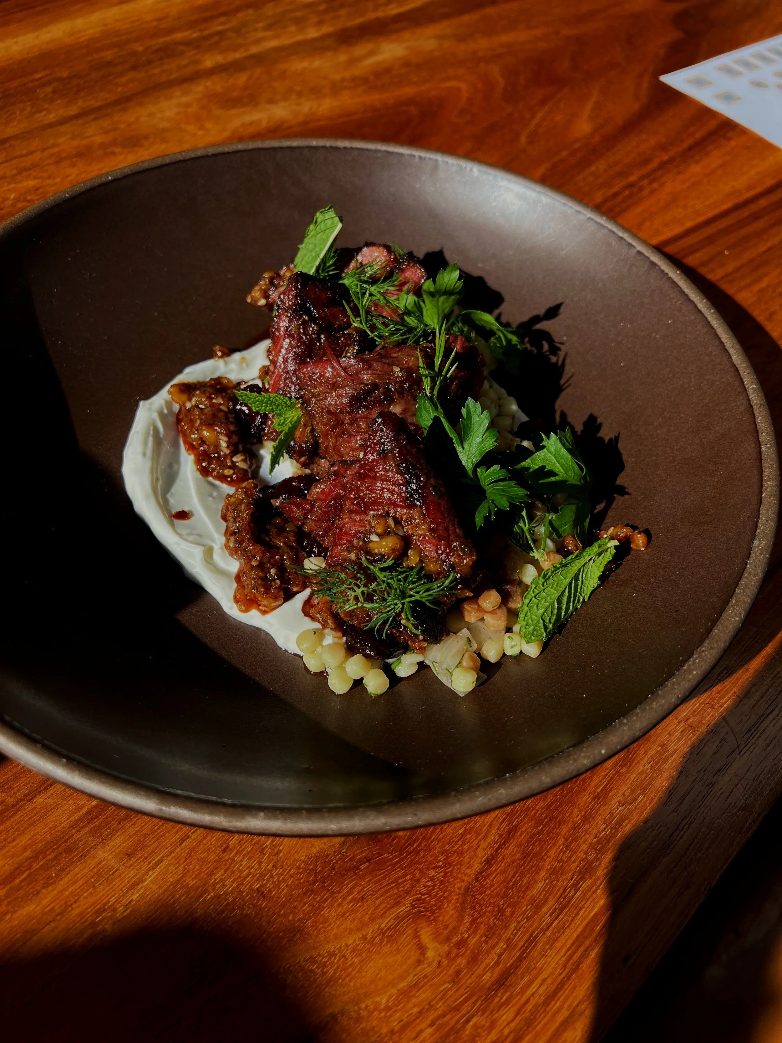 A plate of grilled lamb belly, couscous salad, labneh, and torn fresh herbs.