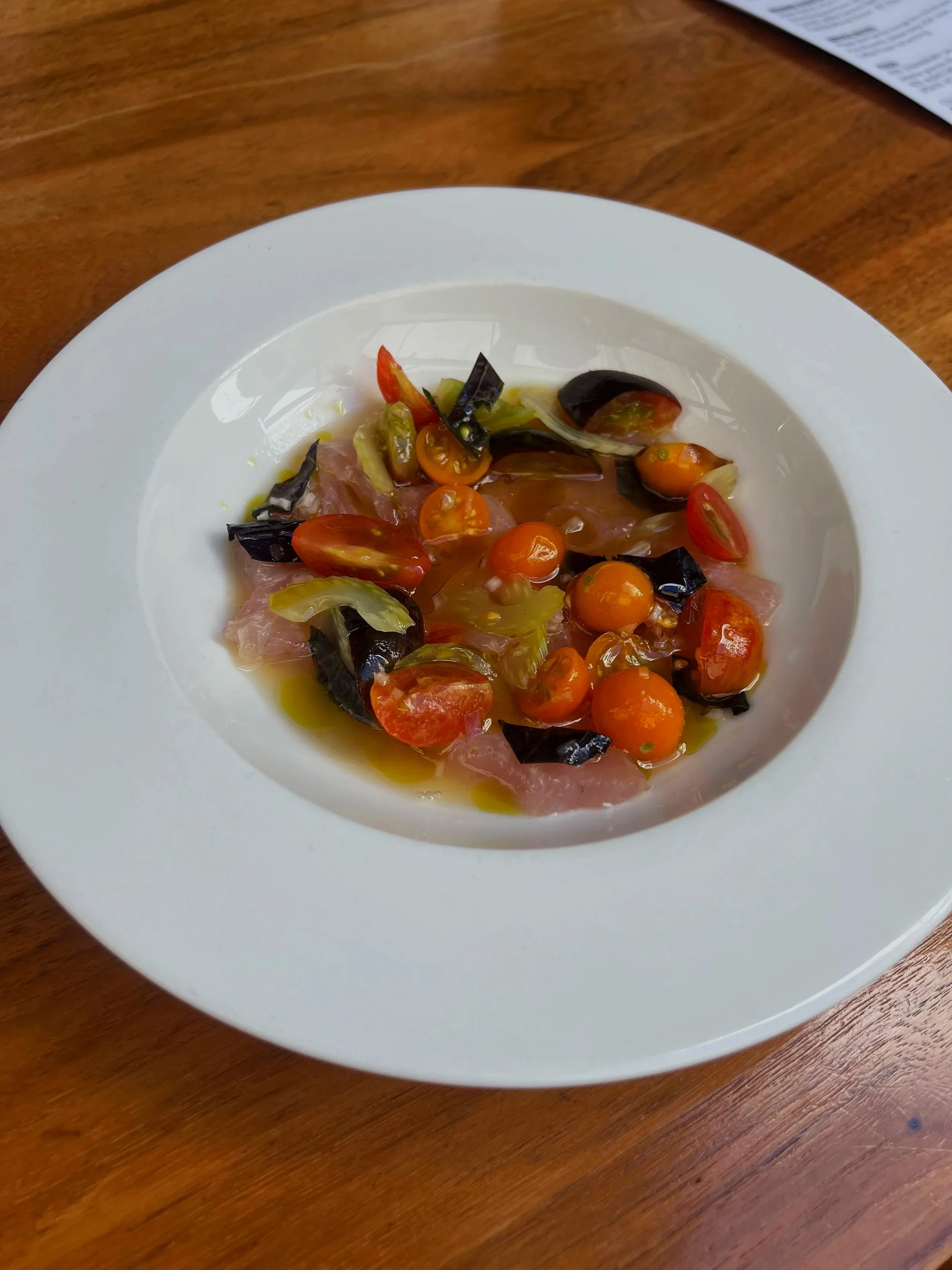 A white bowl containing a tuna crudo with sungold tomatoes, tomato water ponzu, purple shiso, and pickled celery.