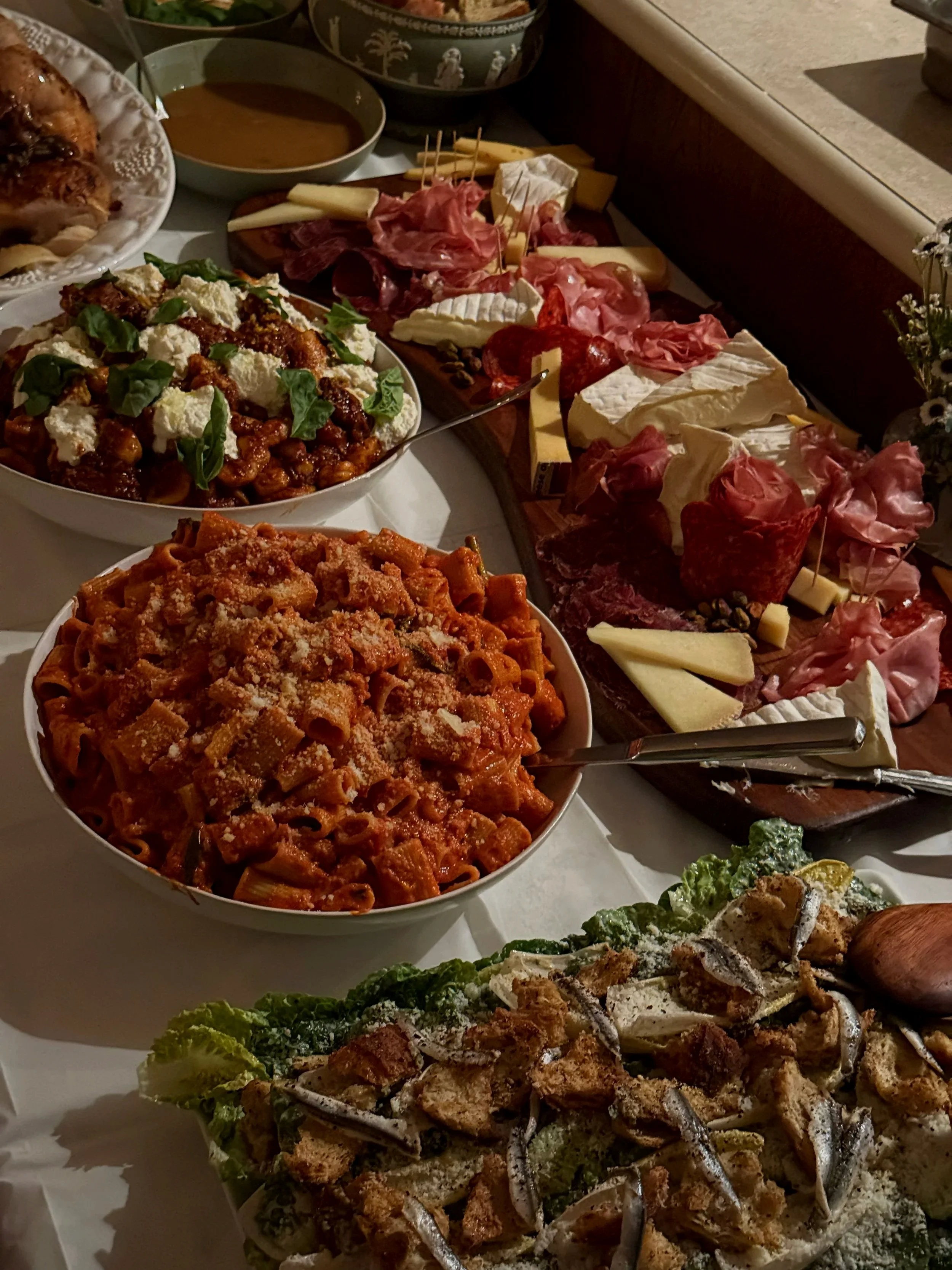 A table with a spread of food including a white anchovy Caesar salad, sausage and ricotta gnocchi, rigatoni amatricana, roasted chicken piccata, and charcuterie.