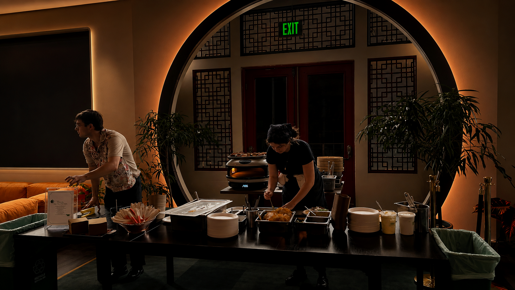 A buffet setup with two staff members preparing food in a restaurant, featuring a large black table with plates, bowls, and food items, with a decorative round window, potted plants, and warm ambient lighting in the background.