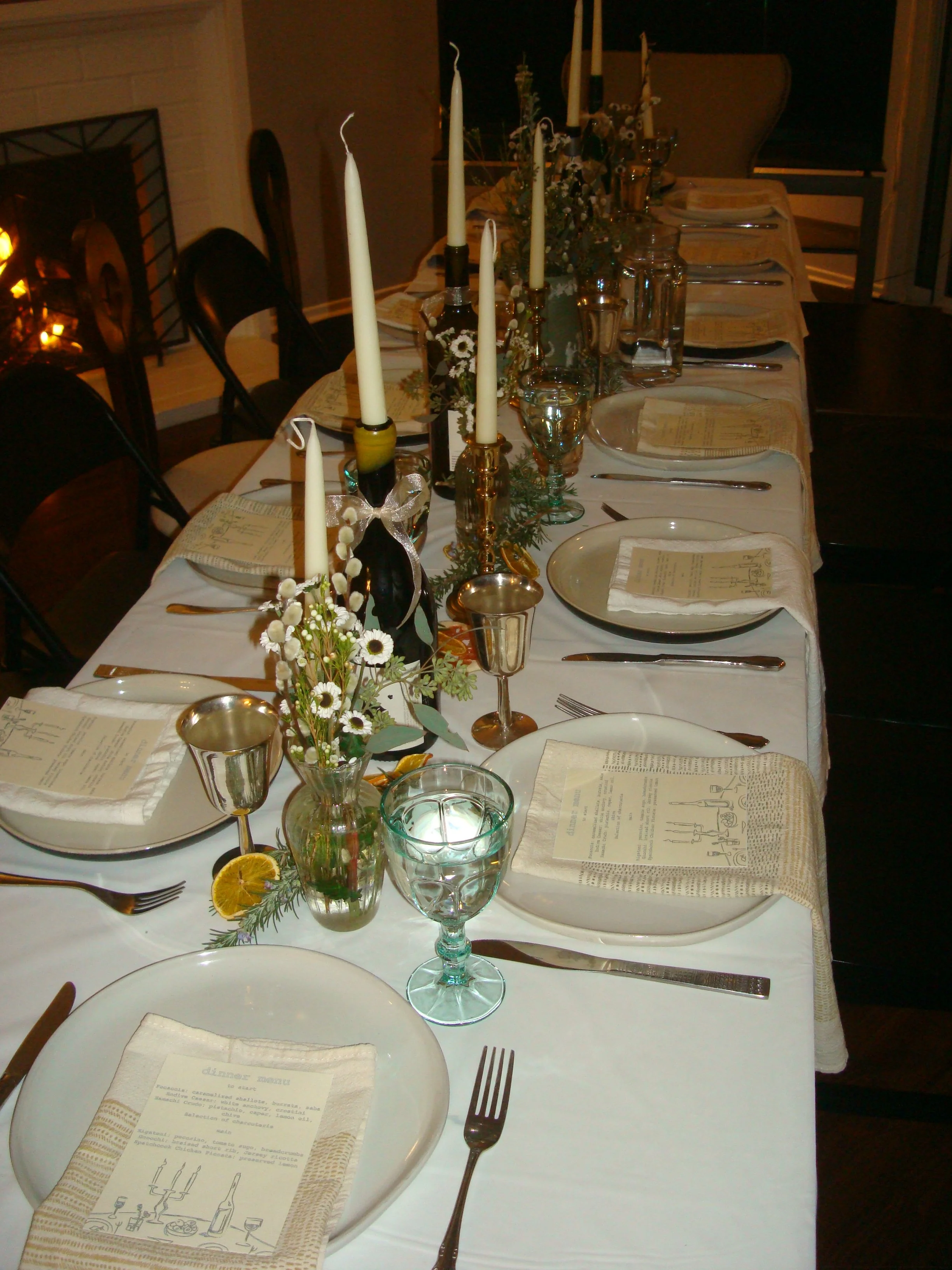 A long dining table set for a meal with plates, silverware, napkins, glassware, and floral arrangements with candles in vases. The setting appears elegant and prepared for a formal dinner event.