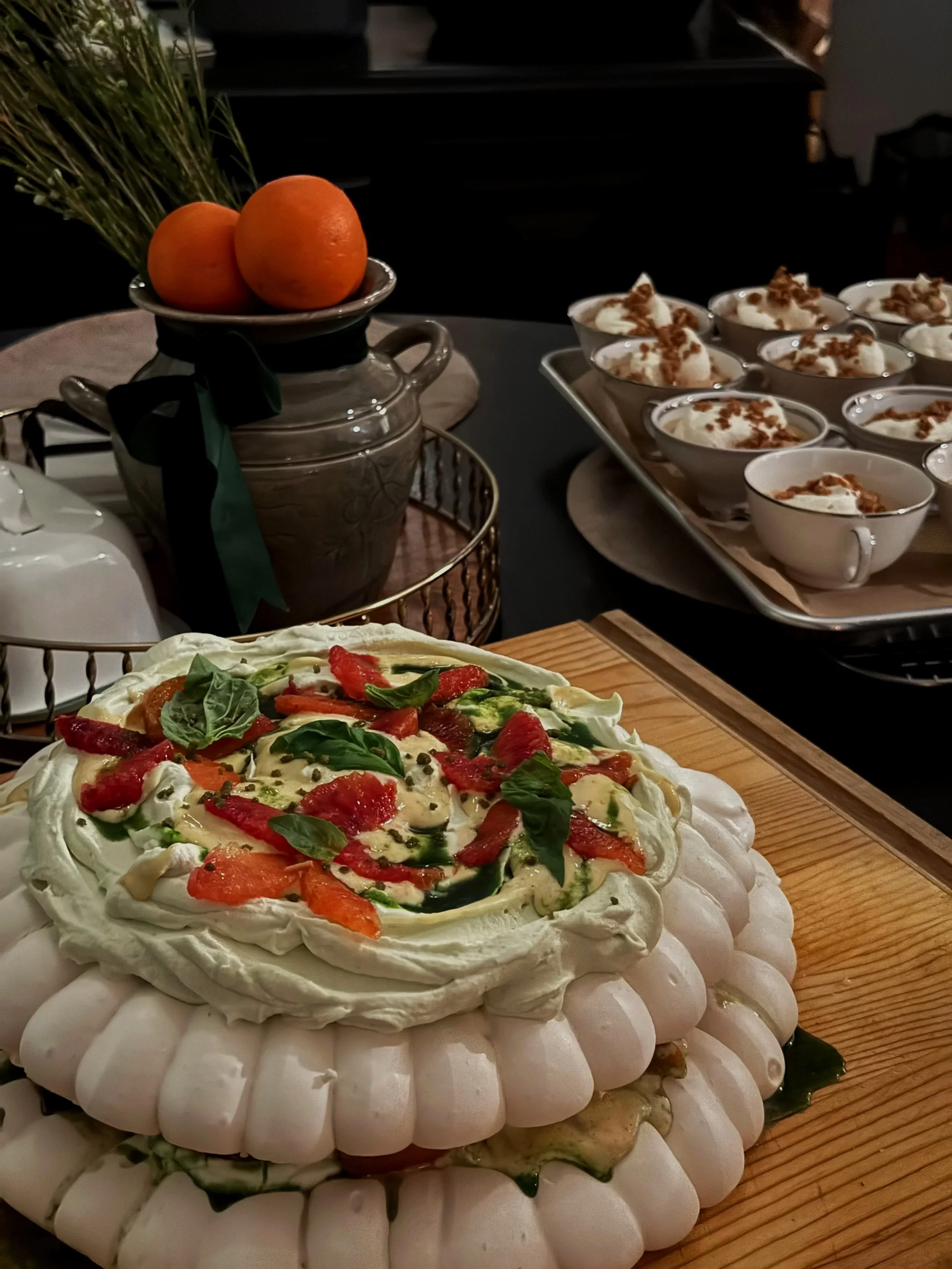 A pavlova topped with basil cream and winter citrus. In the background, there are butterscotch pot de creme.