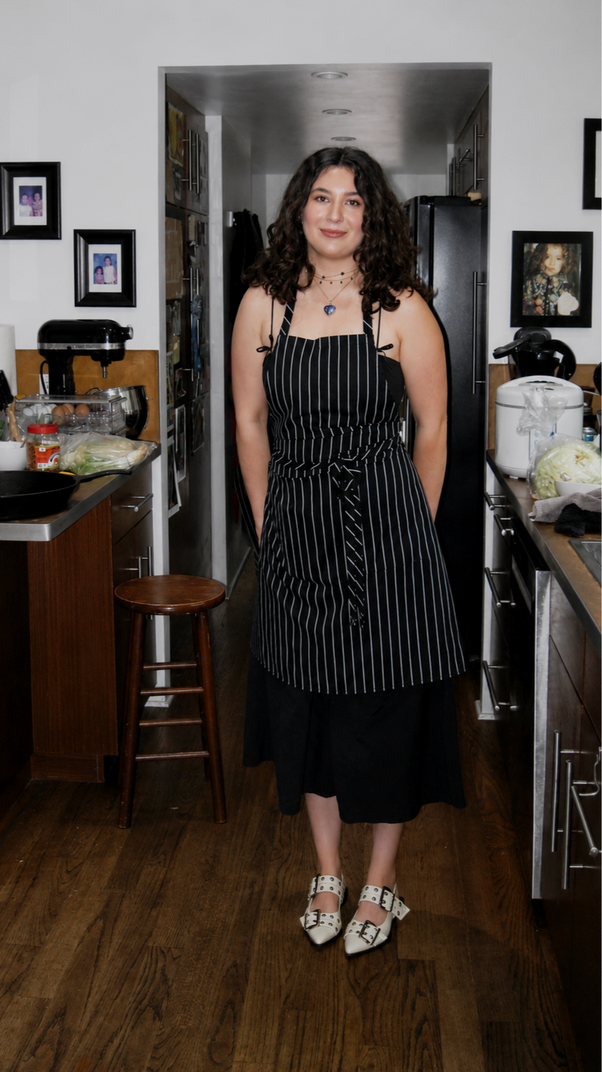 A woman standing in a kitchen, wearing a black and white pinstriped apron over a black dress and white shoes, smiling at the camera.