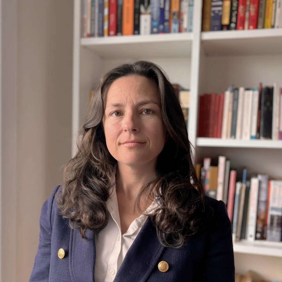 A woman with long brown hair wearing a navy blazer and white shirt, standing in front of a bookshelf filled with books.