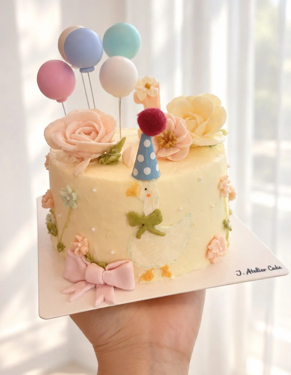 Whimsical Korean-style birthday cake with hand-piped buttercream flowers, a cute goose design, pastel balloons, pink bow, and playful party hat.